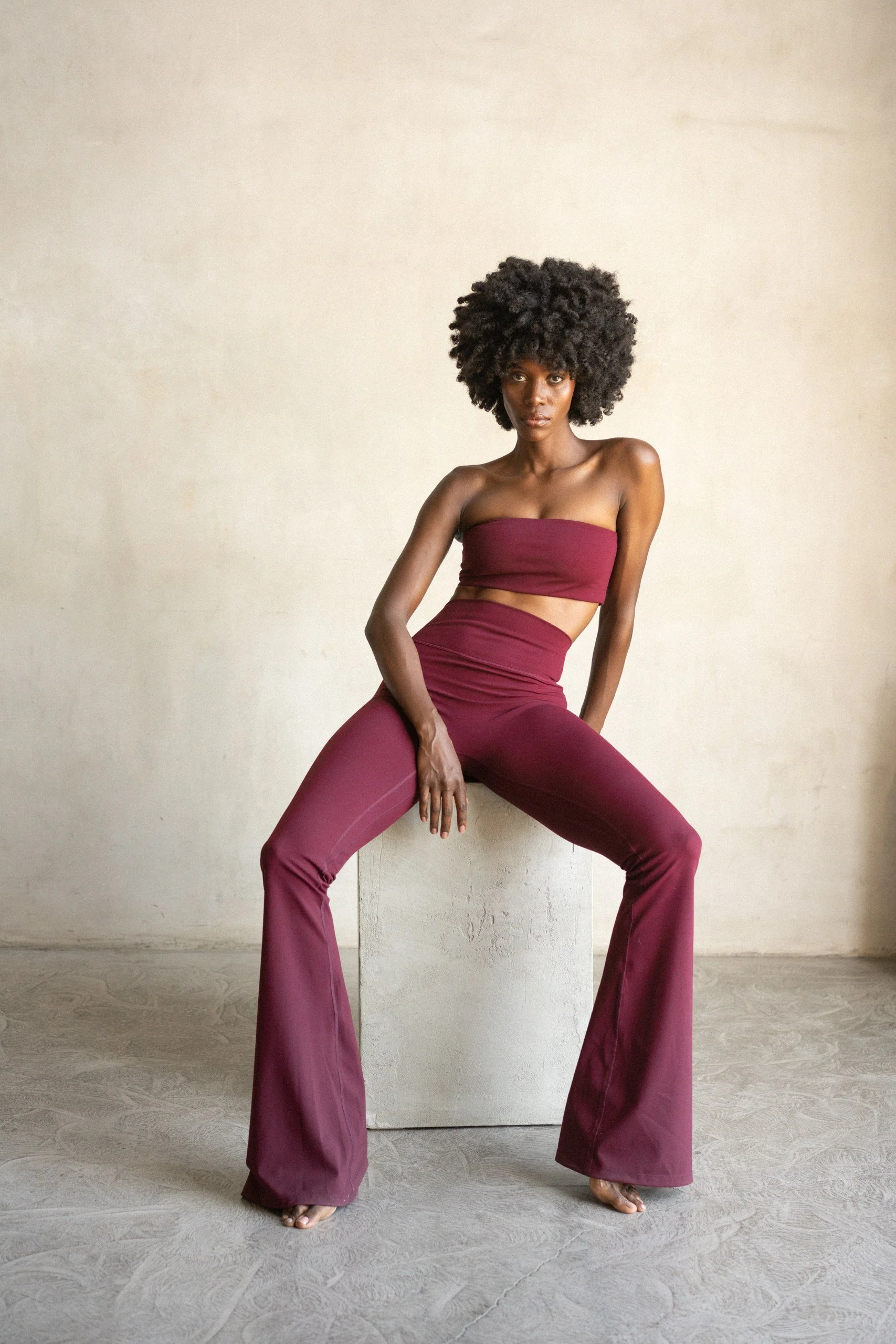 A woman with dark skin and natural curly hair is sitting against a plain neutral wall and floor. She is wearing a strapless maroon crop top and wide-leg maroon pants with a slit. She has a serious expression and is barefoot, with her legs slightly ap