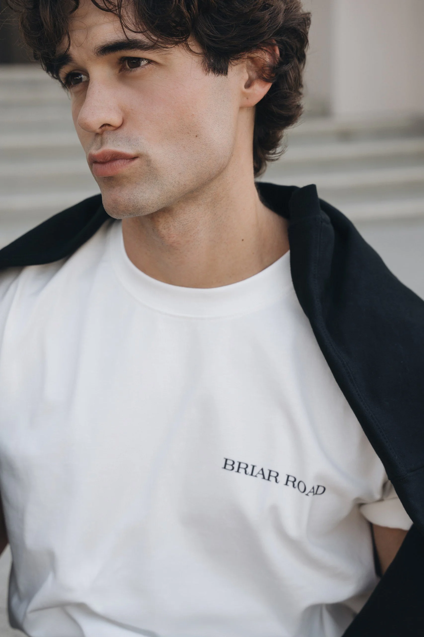 Close-up of a young man with dark, wavy hair, wearing a white T-shirt with the text "BRIAR ROAD" and a black jacket draped over his shoulders, standing outdoors in front of a set of stairs.