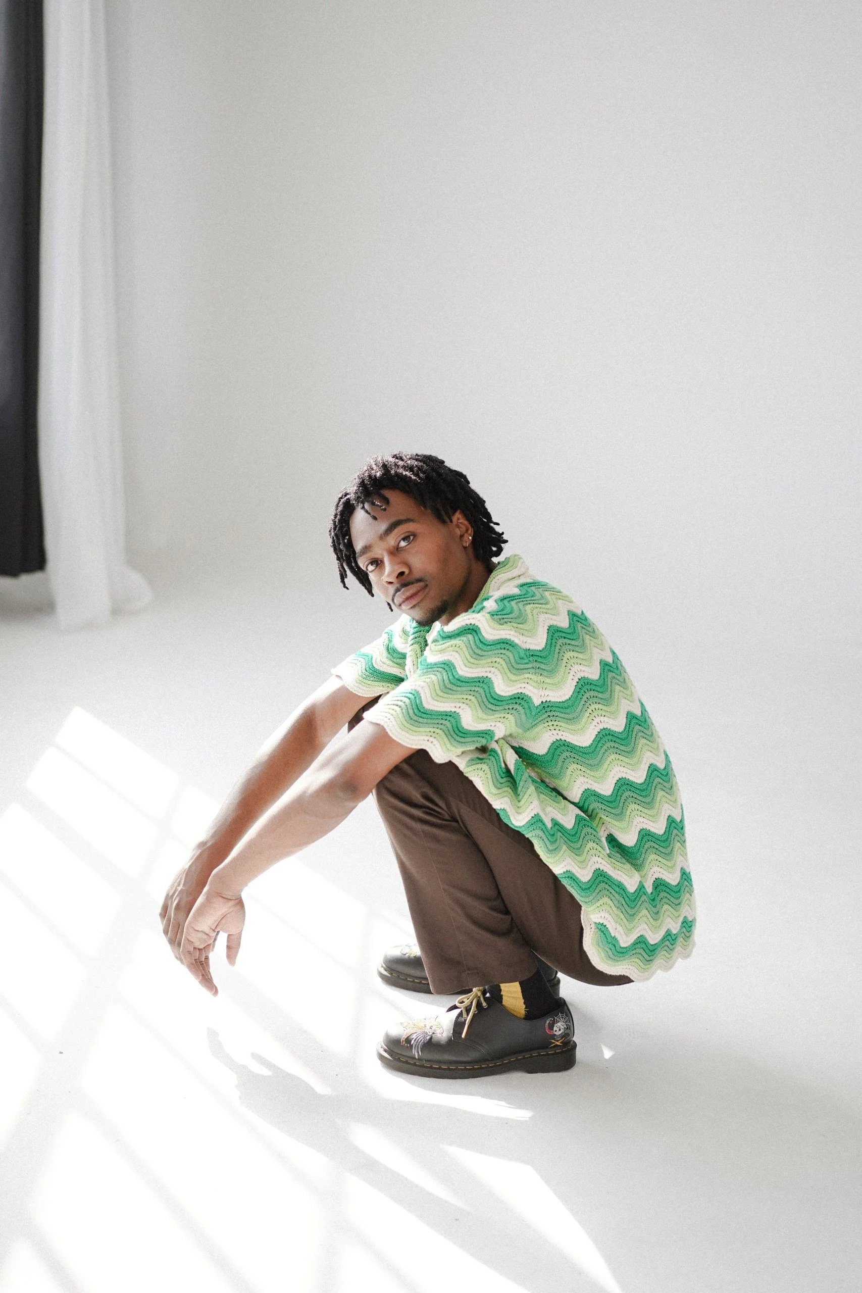 A young man with dreadlocks crouching in a white room with sunlight streaming in through large windows, wearing a green, white, and pink zigzag patterned sweater, brown pants, and black boots.