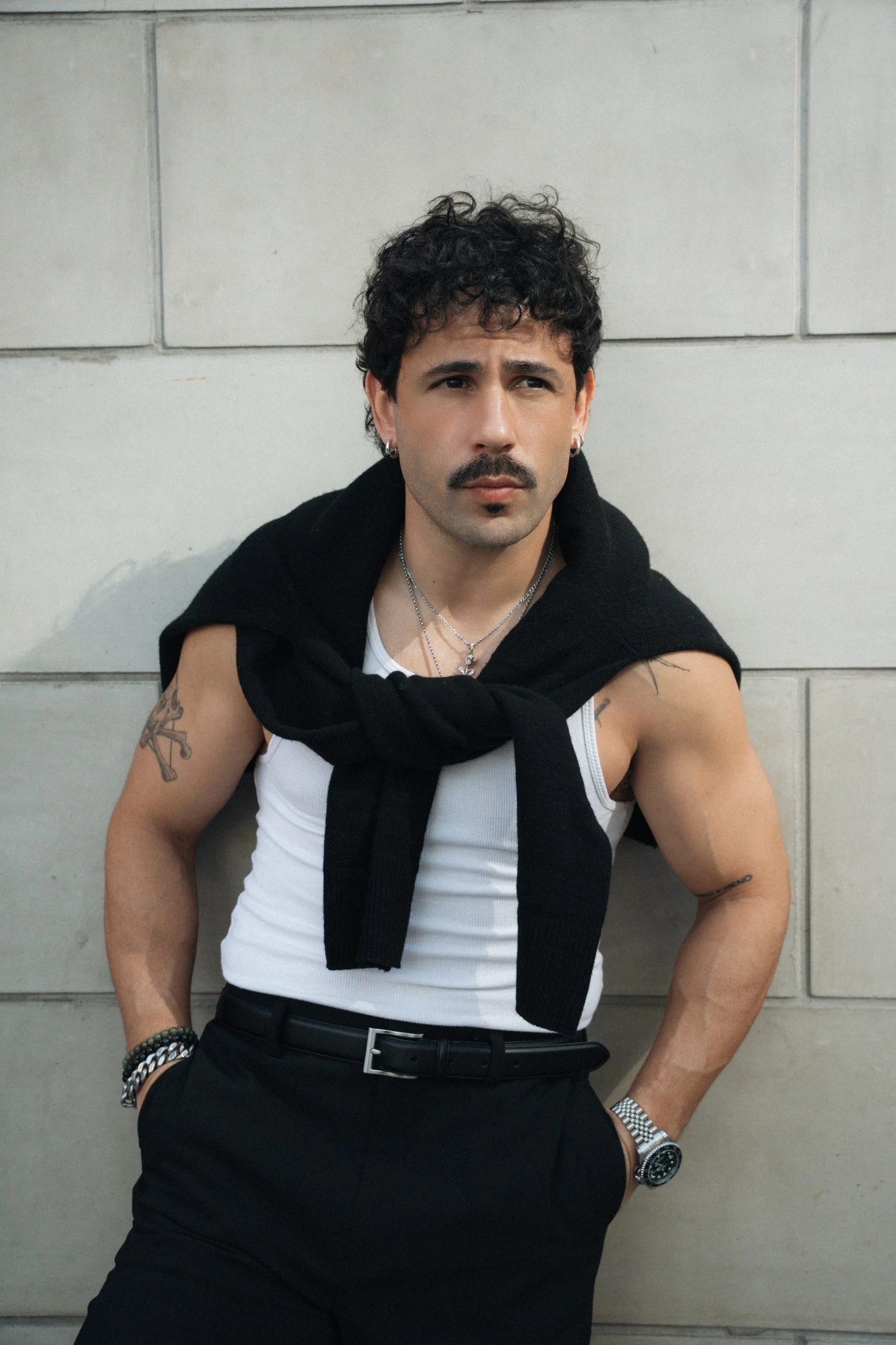 A man with dark curly hair, a mustache, and jewelry, wearing a white tank top with a black sweater tied around his shoulders, standing against a gray tiled wall.