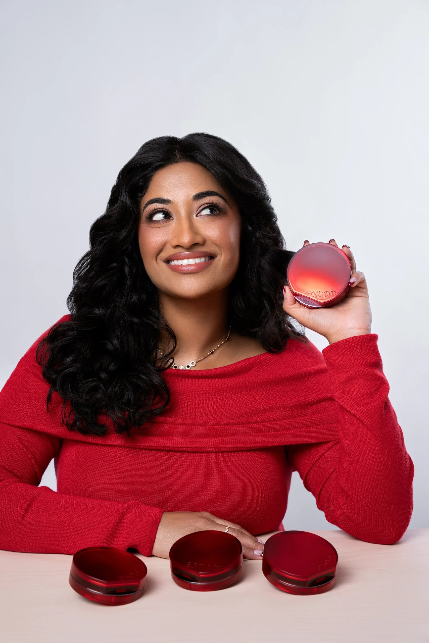 A woman with long, curly black hair smiling and holding a red, round container near her face, with red containers on a light-colored table in front of her.