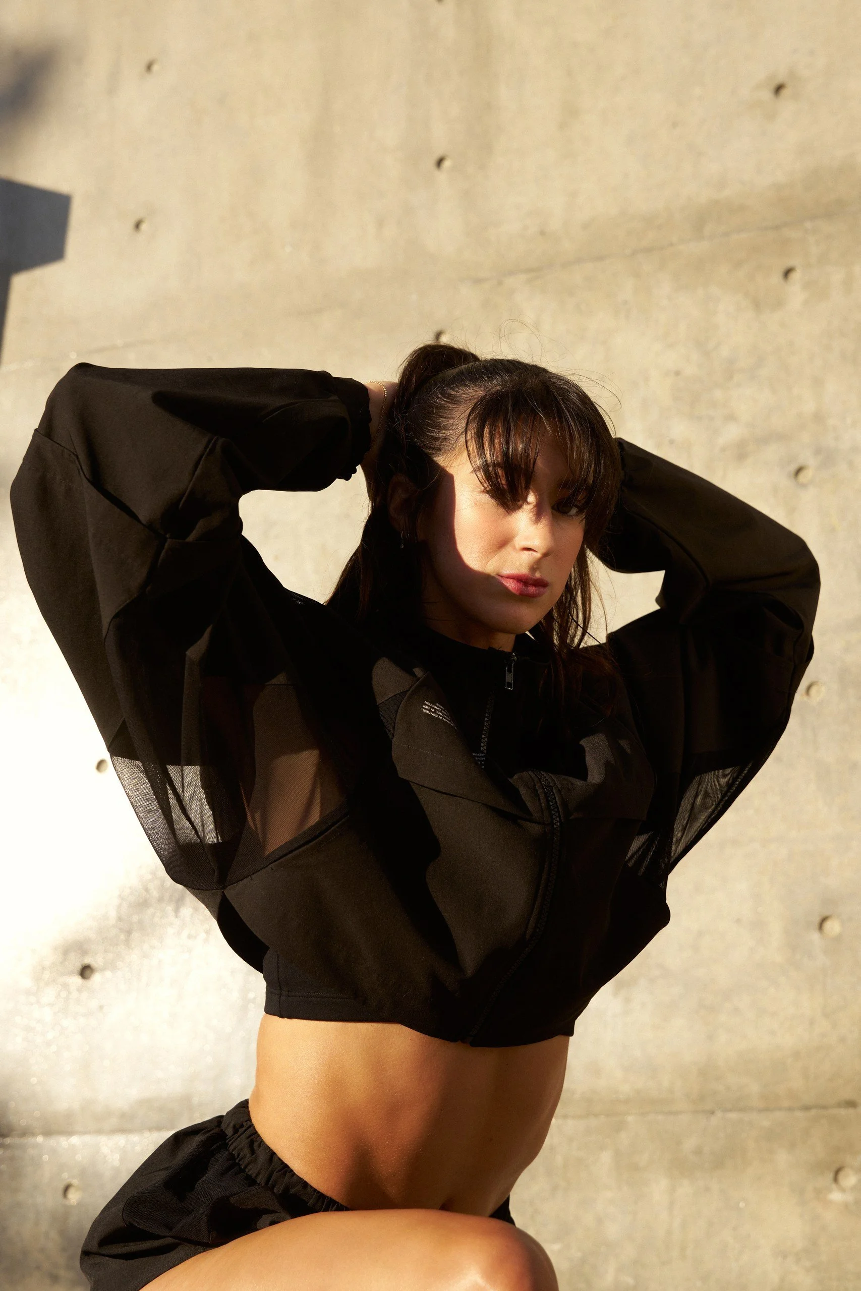 A woman with dark hair in a ponytail wearing a black jacket and black shorts, standing against a concrete wall with her arms raised and hands behind her head.