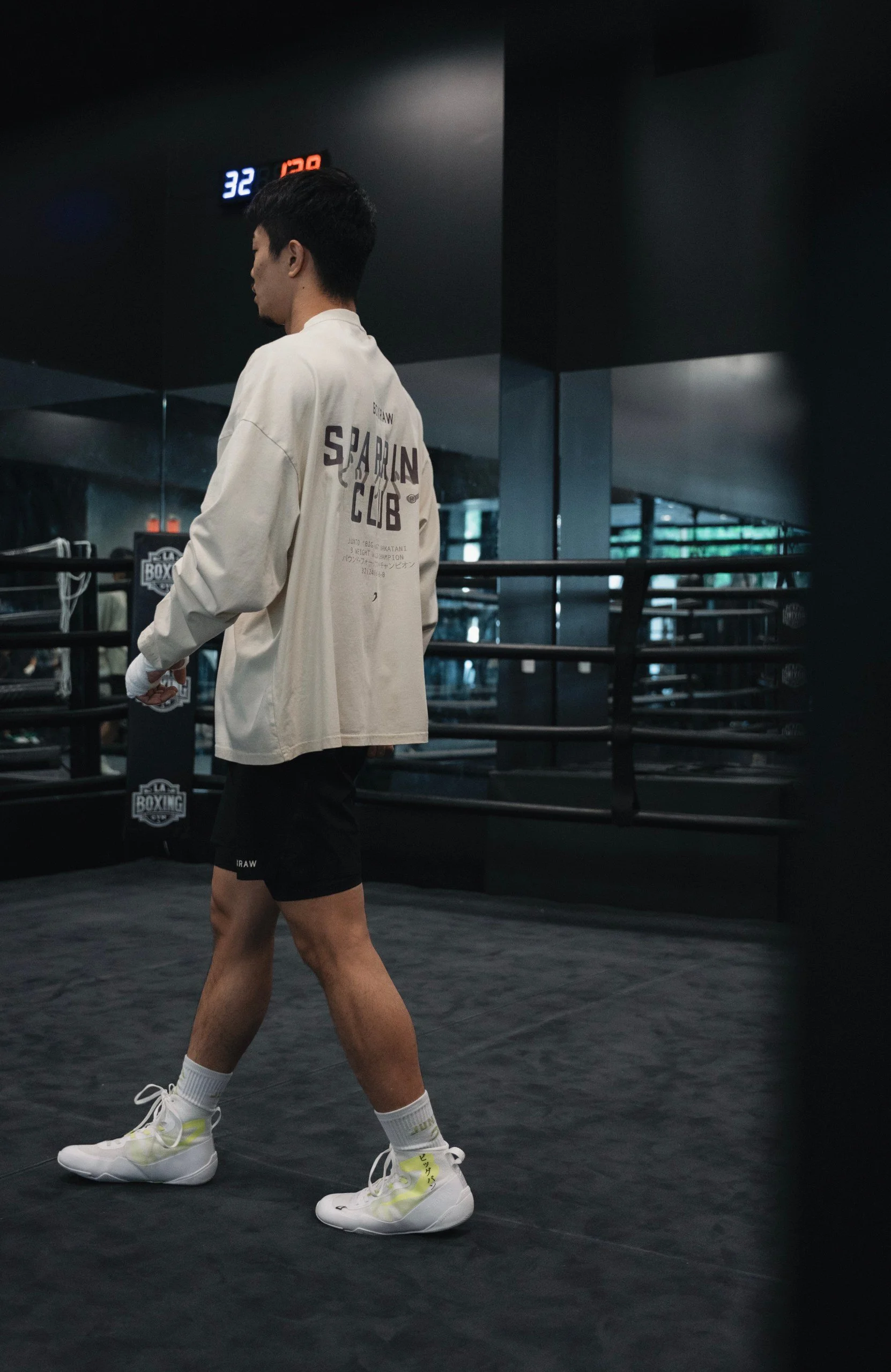 A man walking inside a boxing gym, wearing beige sweatshirt with 'Sparlin Club' on the back, black shorts, white socks, and white sneakers with yellow accents, in front of a boxing ring.