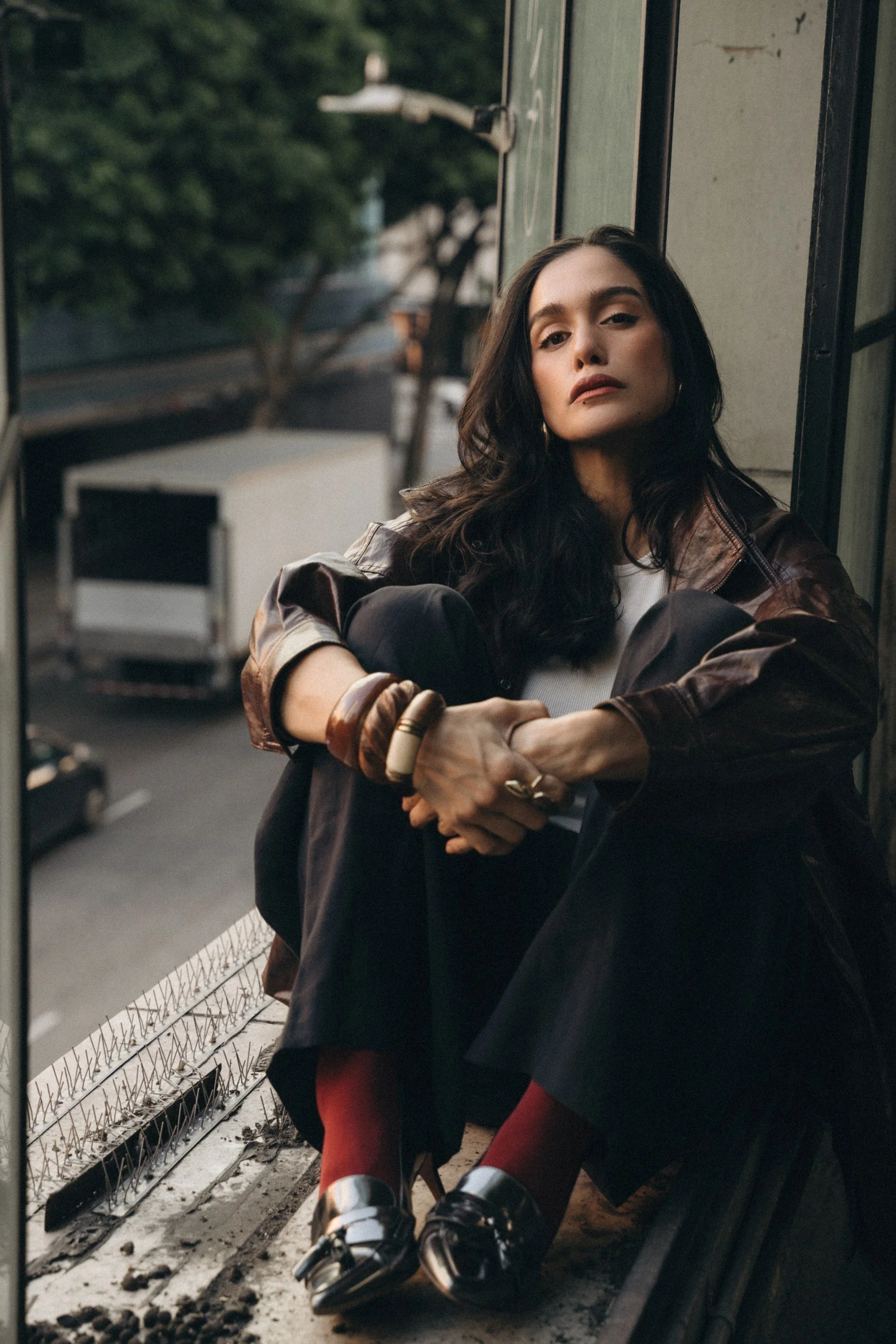 A woman with dark hair and makeup sitting on a city bus or train platform leaning against a wall, wearing a brown leather jacket, black pants, and shiny silver shoes with red socks visible.