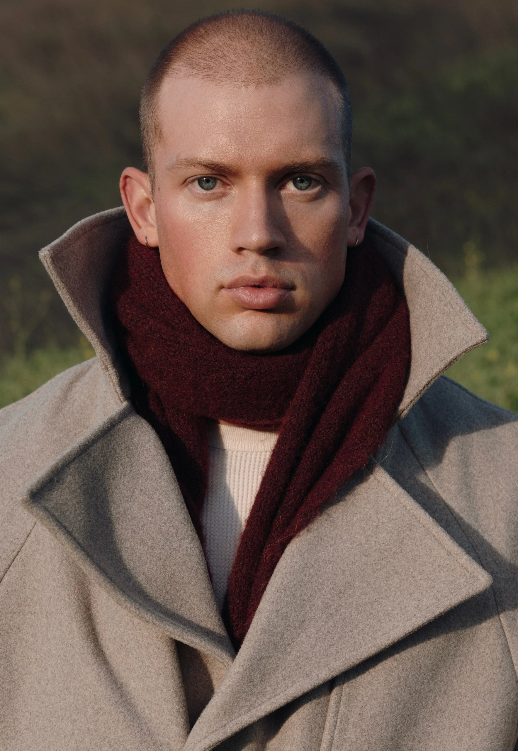 Close-up of a young man with blue eyes, short hair, wearing a beige coat and a burgundy scarf, outdoors with a blurred natural background.