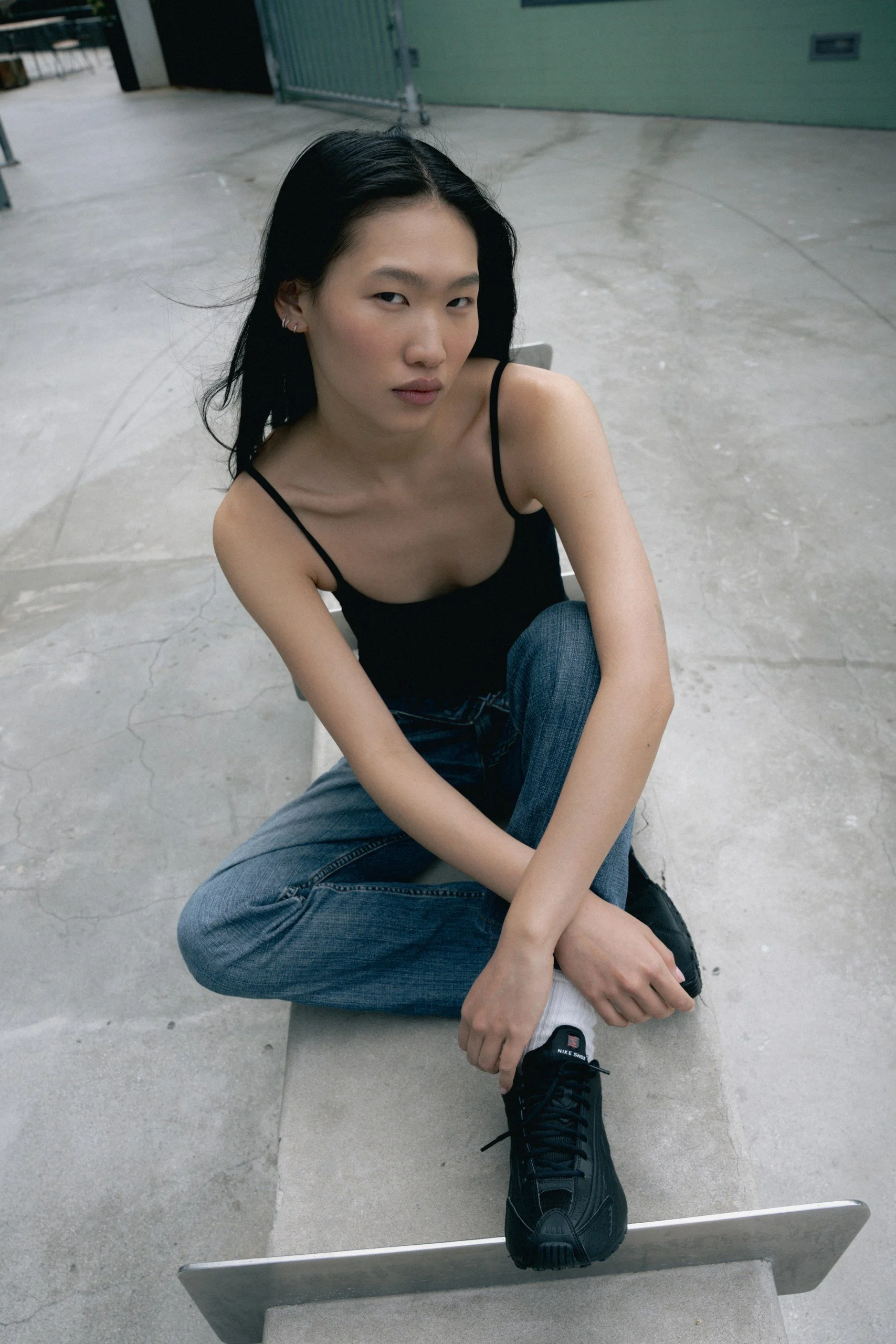 A young woman with long black hair, wearing a black spaghetti strap top, blue jeans, and black Nike sneakers, sitting on a concrete bench in an industrial setting with gray flooring and green metal doors in the background.