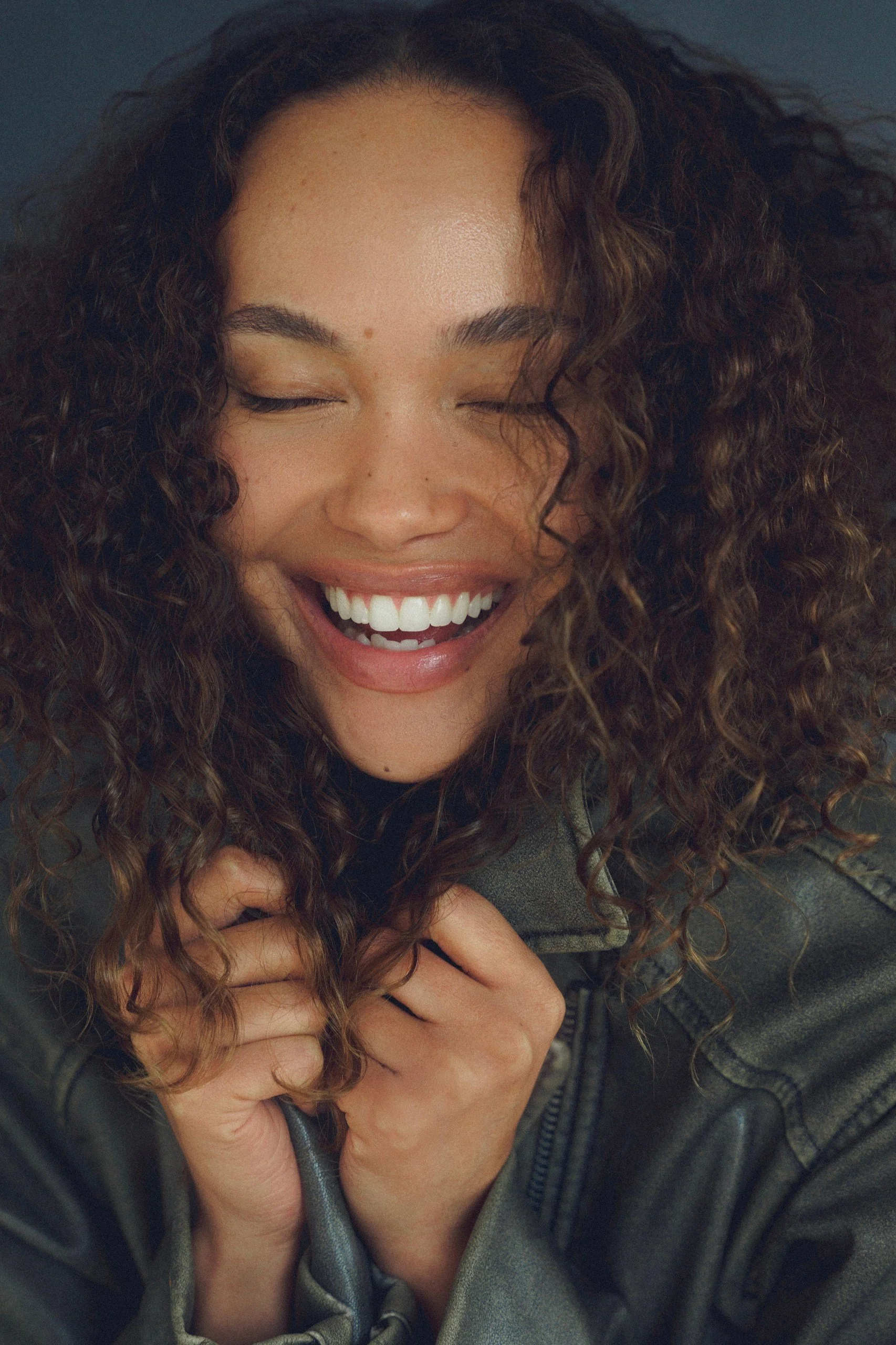 A woman with long, curly hair smiling and laughing with her eyes closed, wearing a grayish-green jacket.
