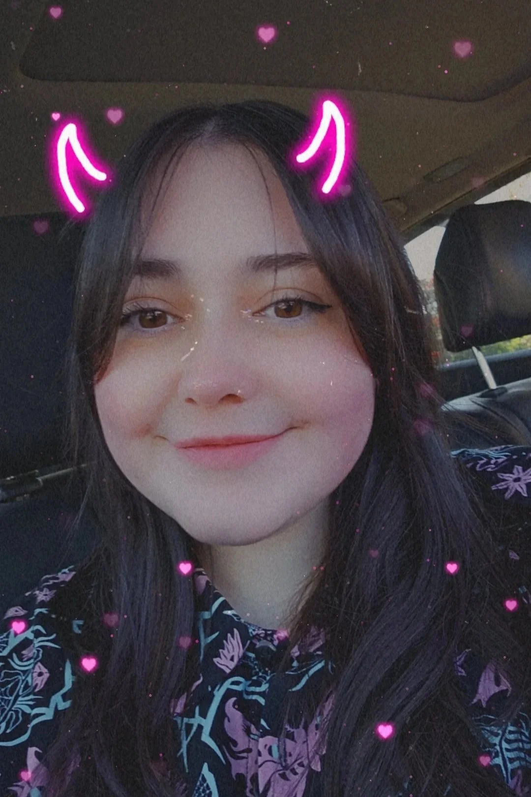 Salem with dark hair and light skin, smiling with a filter that adds pink devil horns and many small pink hearts around her, sitting inside a vehicle.