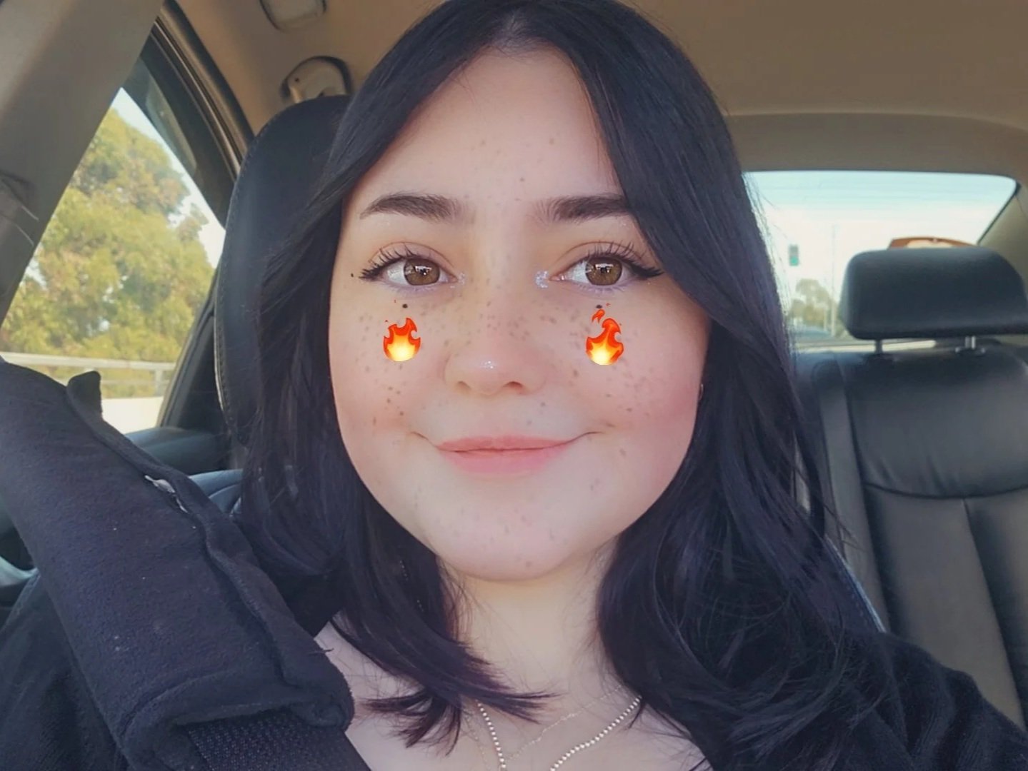 Salem with dark hair and brown eyes smiling inside a car, with filters adding flame icons under her eyes and light makeup.