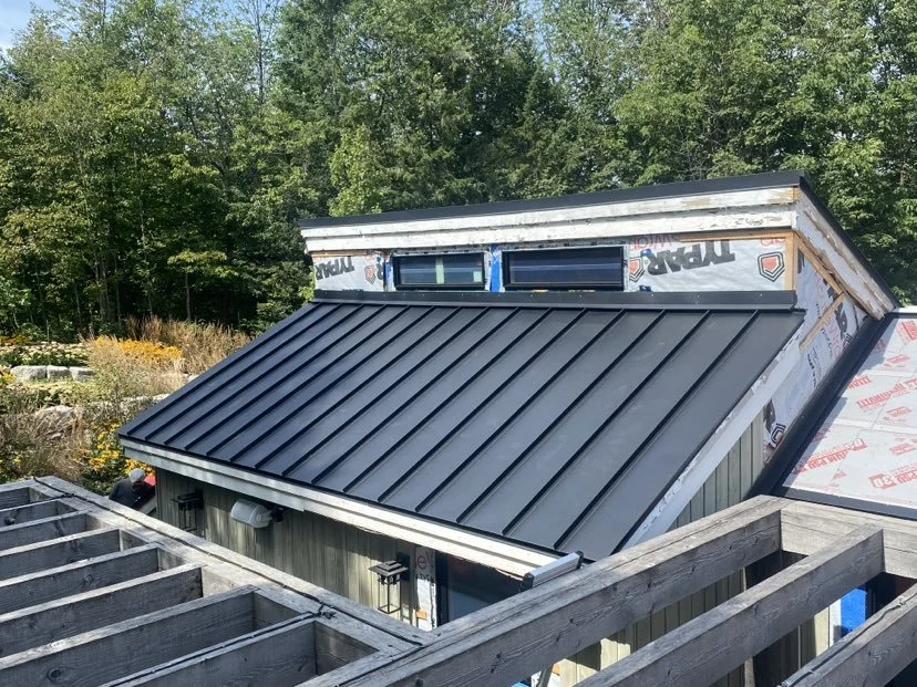 Construction of a new metal roof on a house, with a partially completed window area and surrounding greenery.