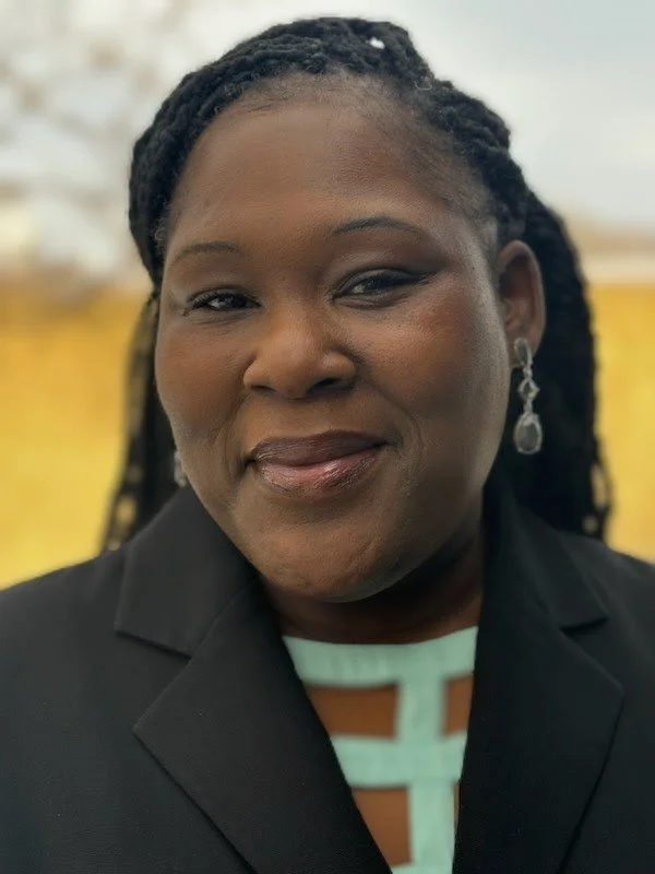 Close-up of an African-American woman with braided hair, smiling, wearing earrings, a black blazer, and a patterned top, against a blurred indoor background.