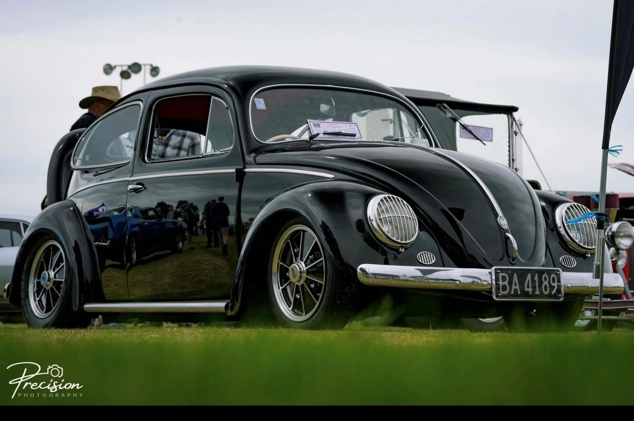 Black vintage Volkswagen Beetle car on display at an outdoor car show, with people in the background and a cloudy sky overhead.