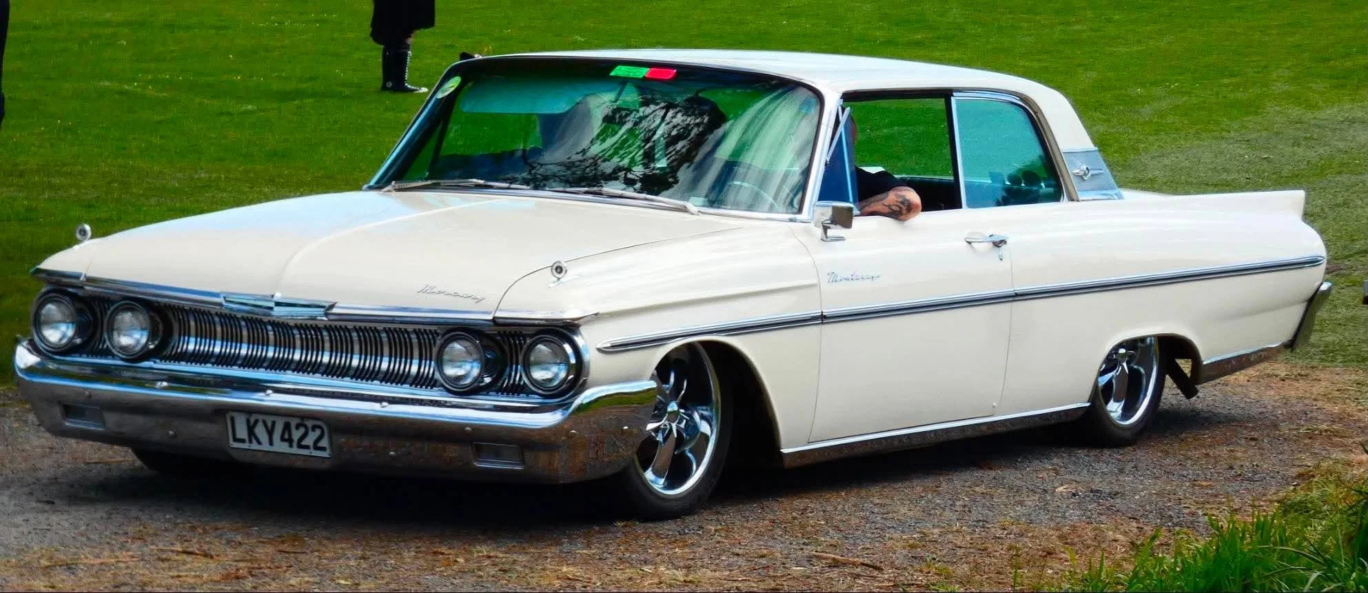 A vintage white Mercury Monarch car parked on a grassy area, with someone inside and another person standing nearby.