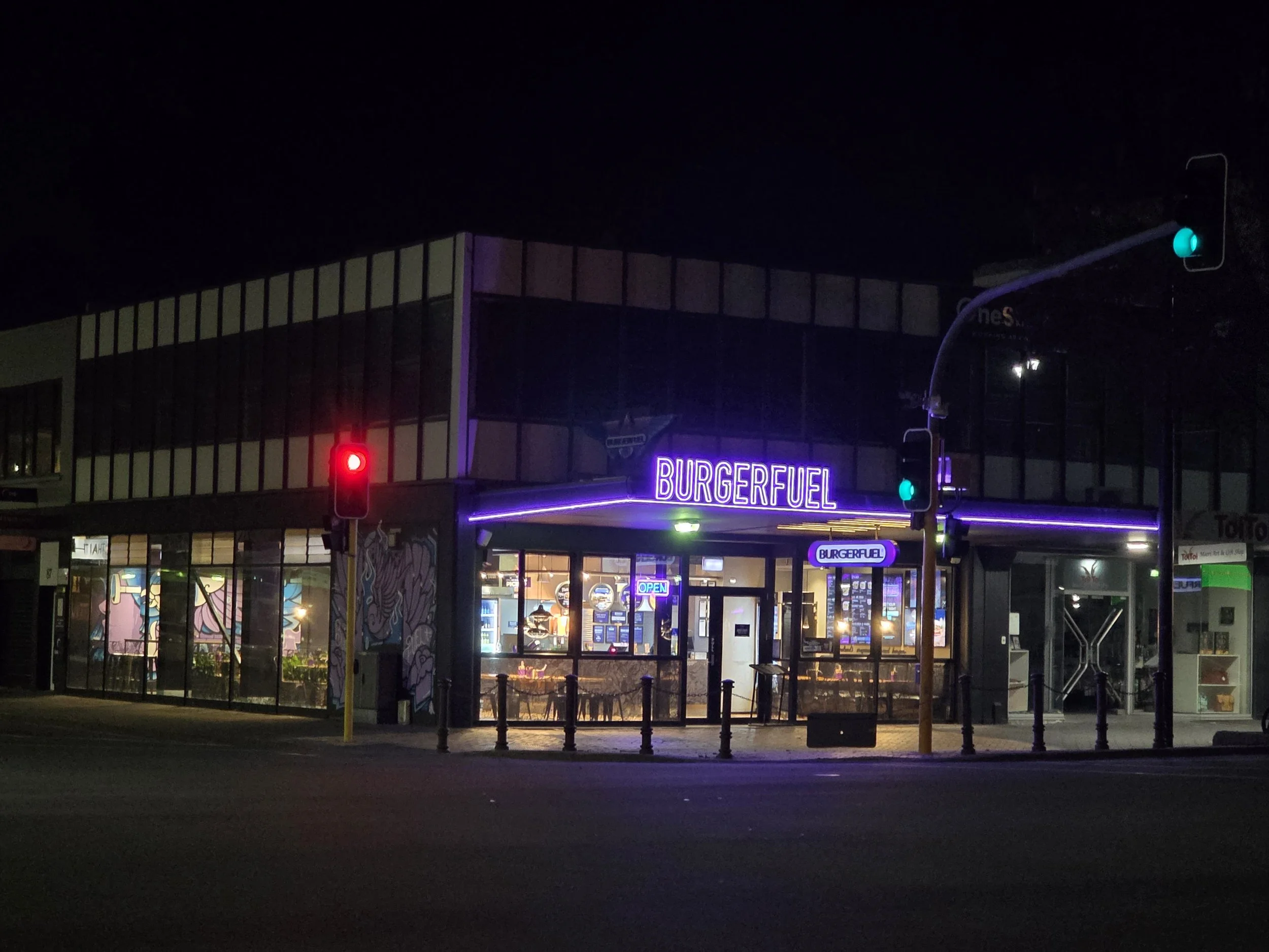 Nighttime exterior of a BurgerFuel restaurant with neon signs and traffic lights.