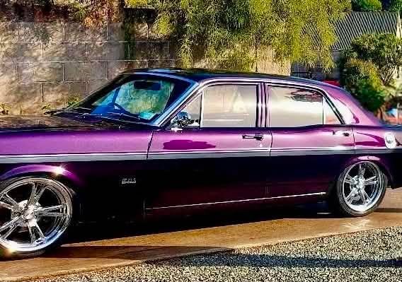 A purple and black vintage car parked on a gravel driveway with trees and a brick wall in the background.