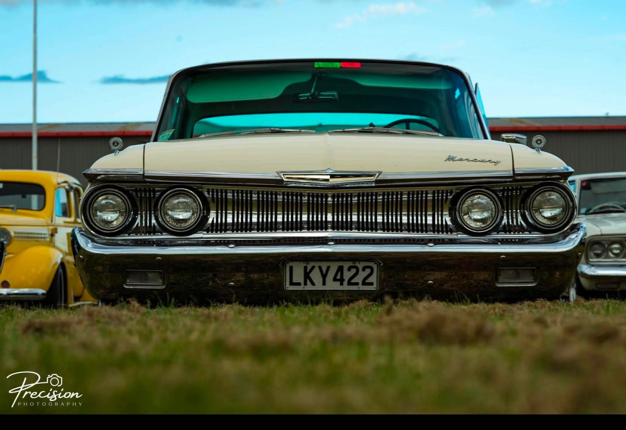 Front view of a vintage cream-colored Mercury car with a black grille, parked among other classic cars at a car show. The Mercury logo is visible on the front, and the car has a clear windshield and dual round headlights.