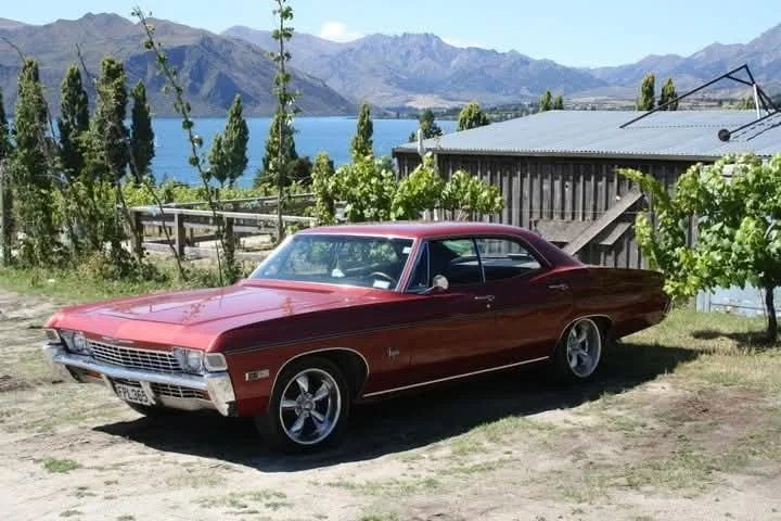 A vintage red Chevrolet muscle car parked on a dirt area near a vineyard with mountains and a lake in the background.