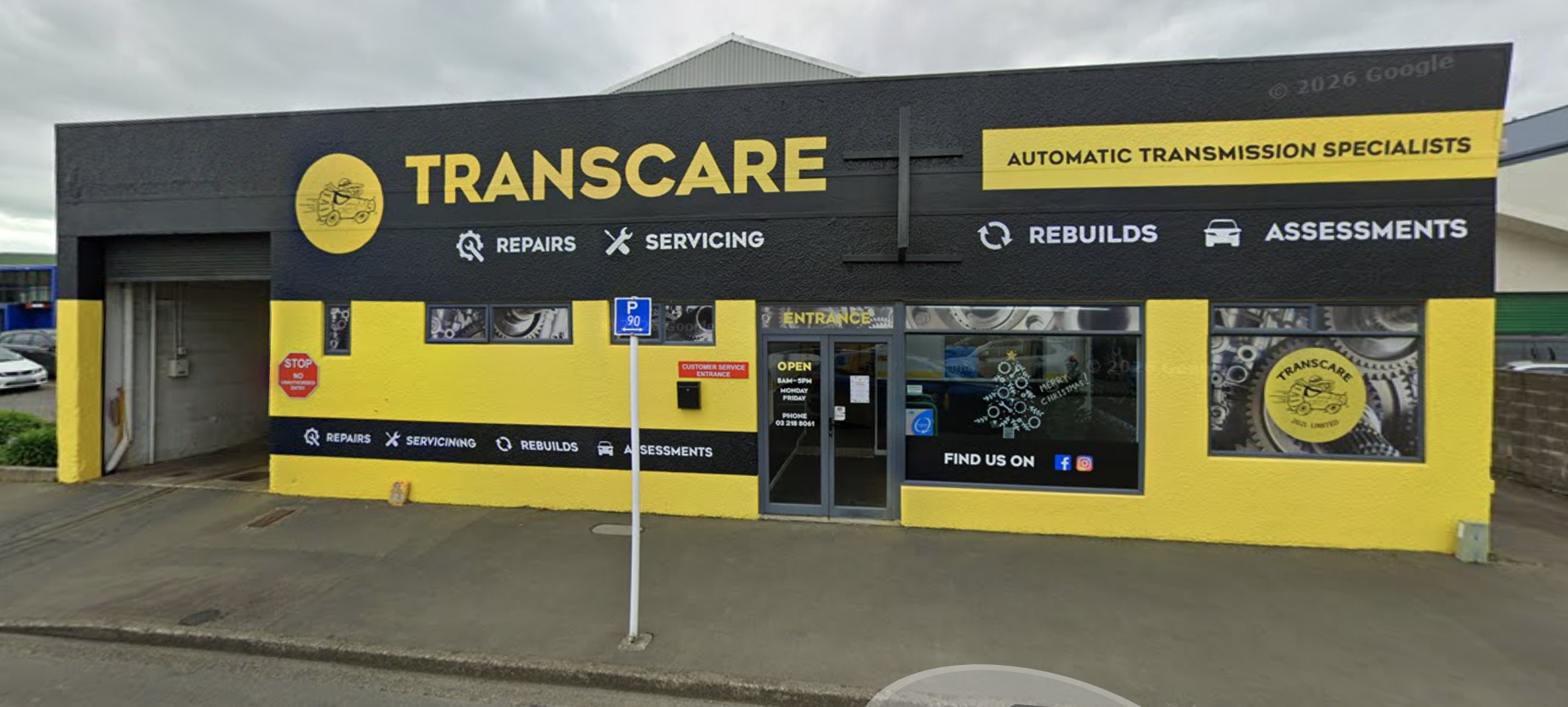 Exterior view of an auto repair shop named Transcare, featuring a black and yellow color scheme, signs indicating services like repairs, servicing, rebuilds, assessments, and an entrance door with signage and social media icons.