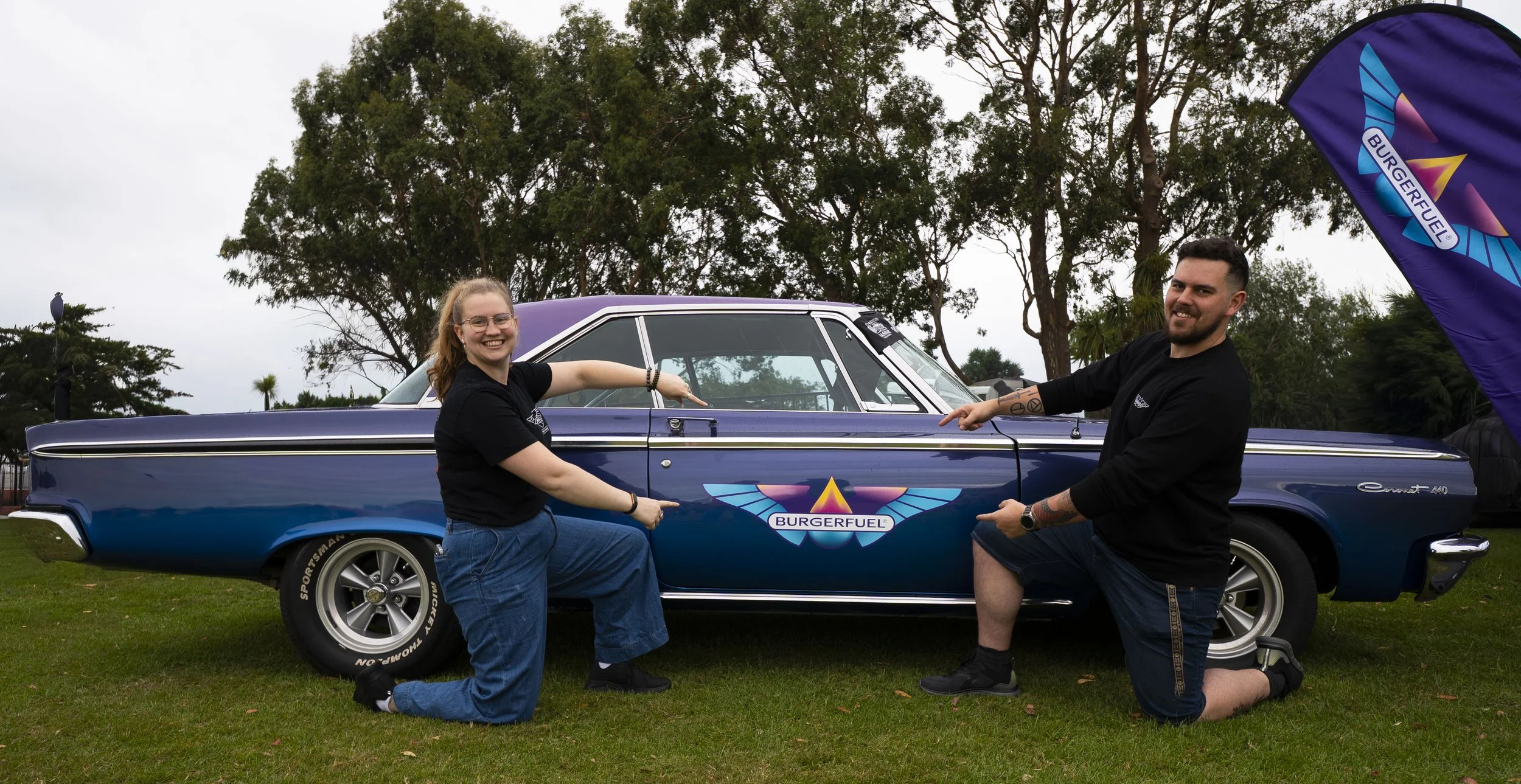 Two people, one woman and one man, kneel in front of a vintage blue car with a BurgerFuel logo on the side, pointing at the logo, outdoors with trees and an overcast sky.