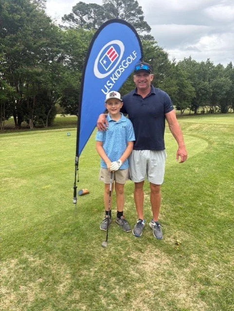 A young boy and an adult man stand together on a golf course, smiling, with their arms around each other. They are near a blue flag that reads "US KIDS GOLF". The boy holds a golf club and wears a baseball cap, while the man wears a cap, a navy blue shirt, and gray shorts. The background shows trees and a cloudy sky.