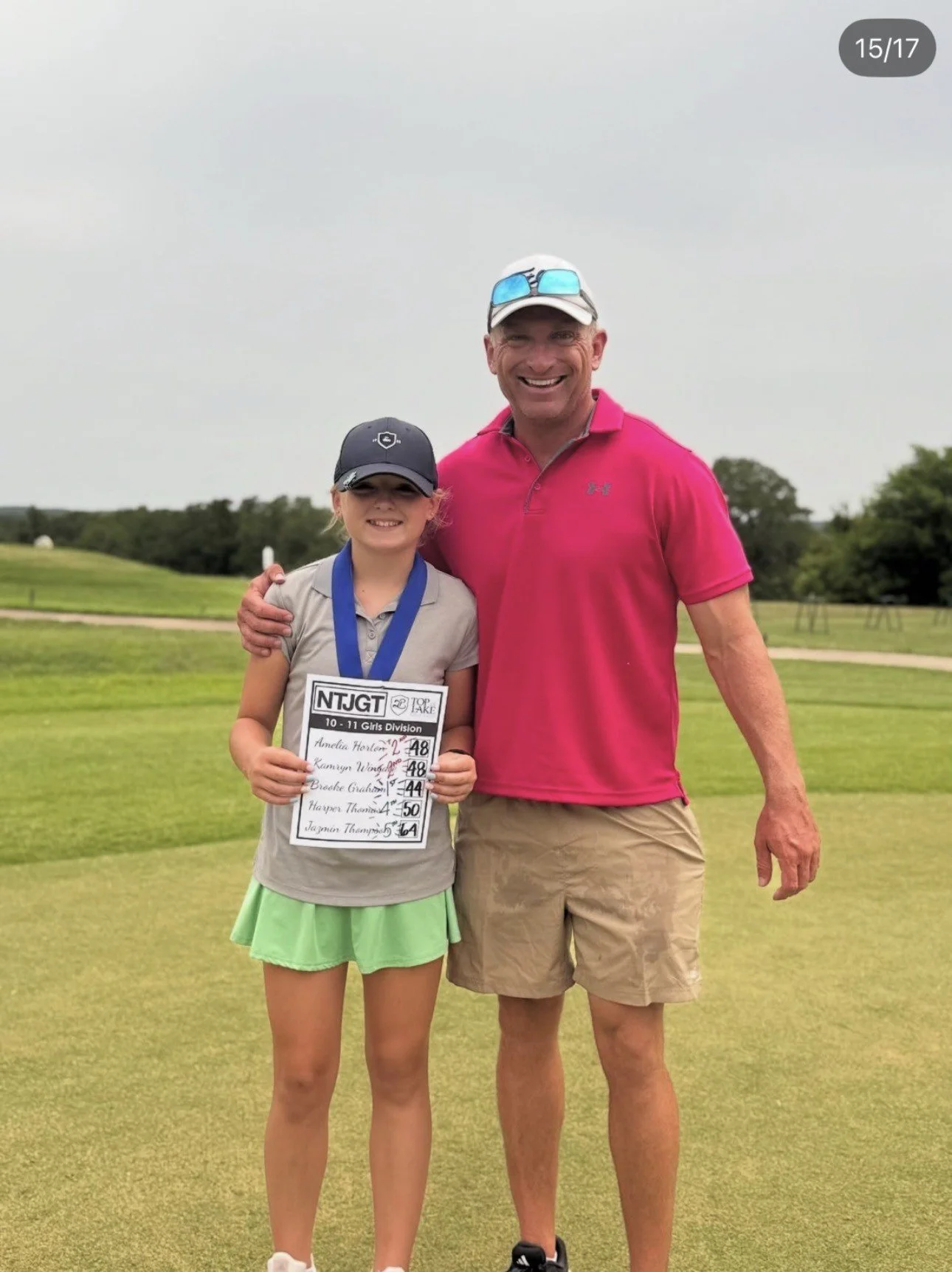 A girl and a man standing on a golf course, smiling for a photo. The girl is holding a golf tournament scorecard. The girl is wearing a black cap, light gray shirt, and a lime green skirt, with a medal around her neck. The man is wearing a pink polo shirt, tan shorts, and a white cap with sunglasses on top.