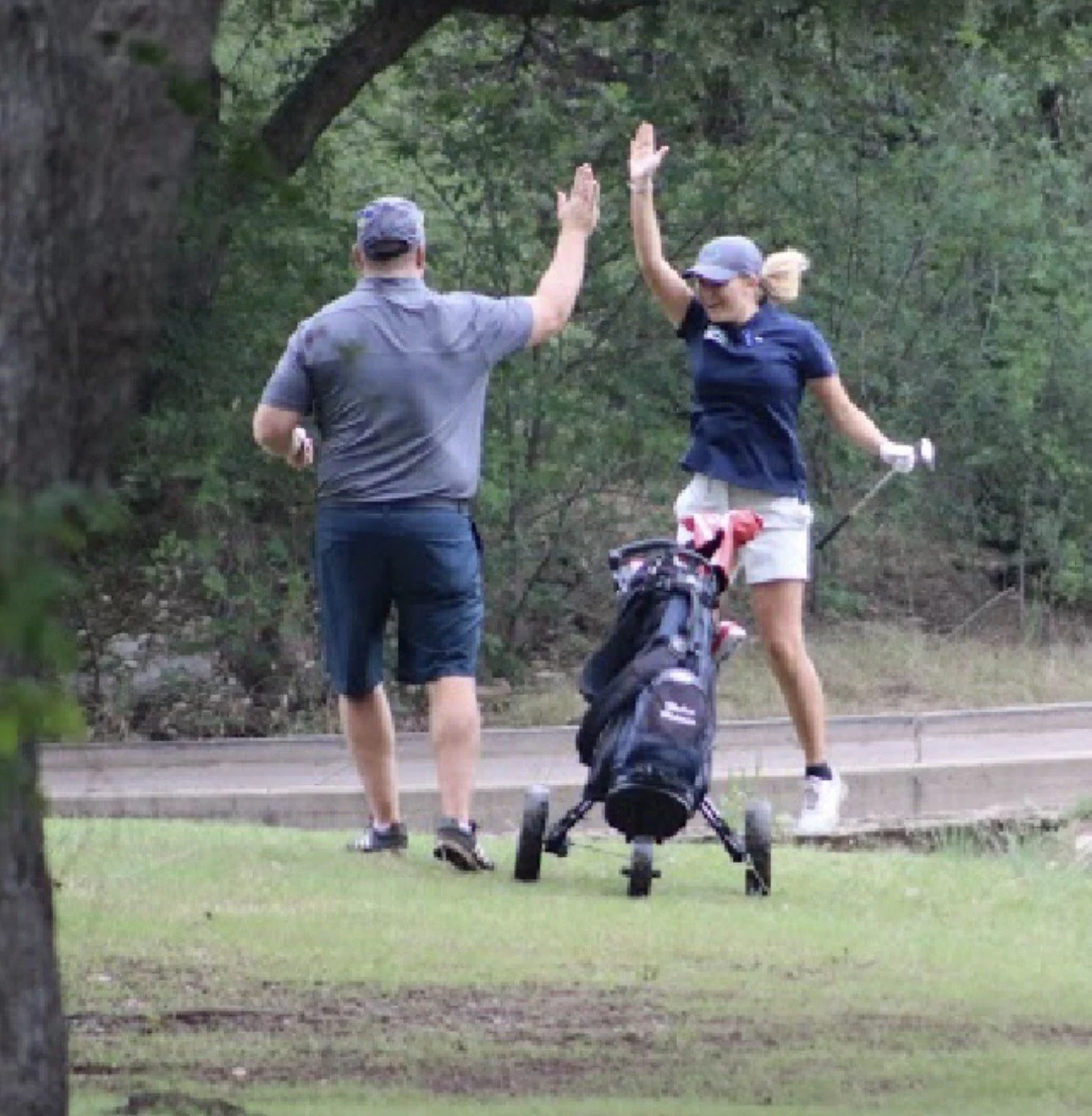 A man and woman on a golf course giving each other a high five, with a golf bag on a pushcart between them.