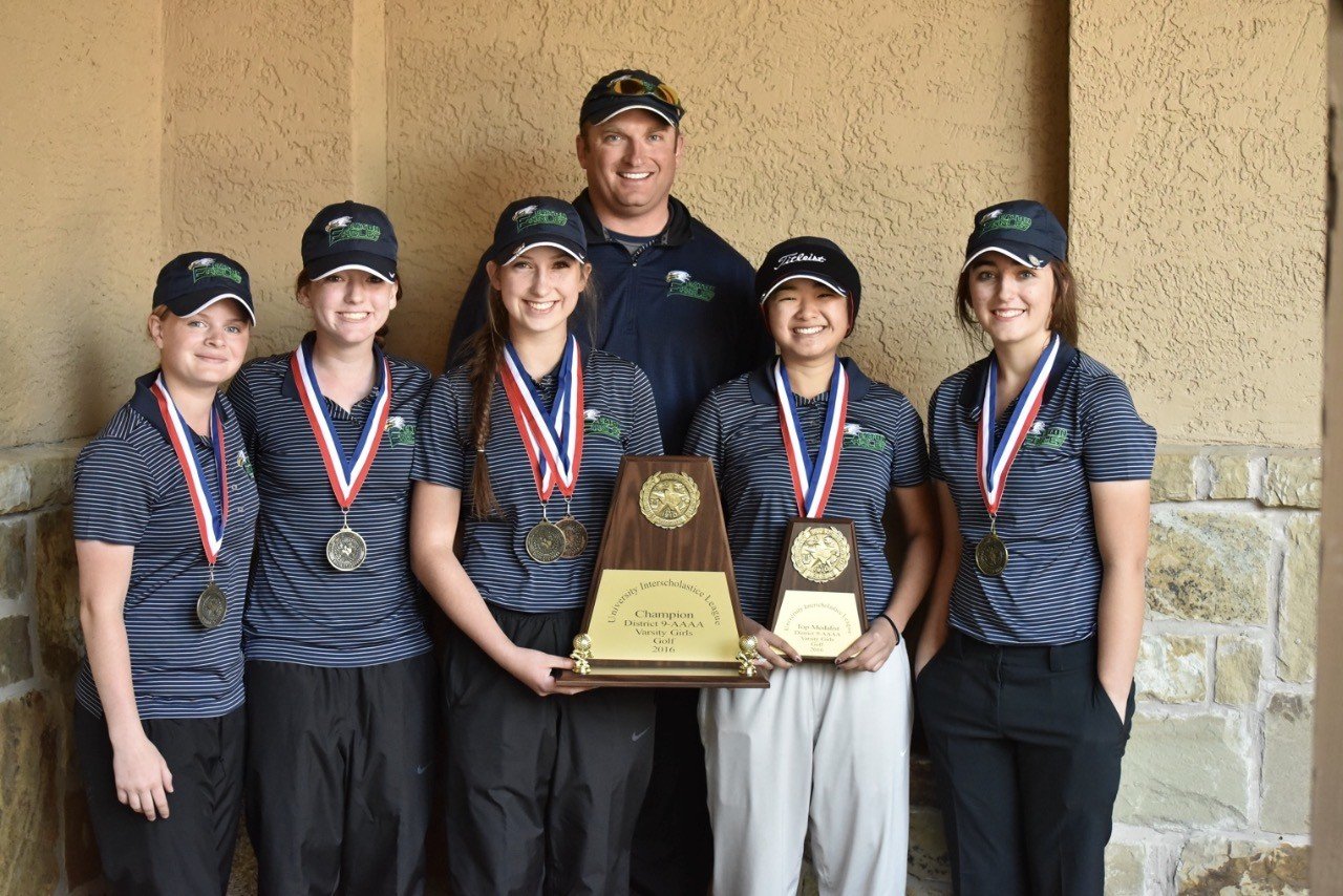Group of six young girls and one man posing with trophies and medals, all dressed in matching golf uniforms and caps, standing indoors against a textured beige wall.