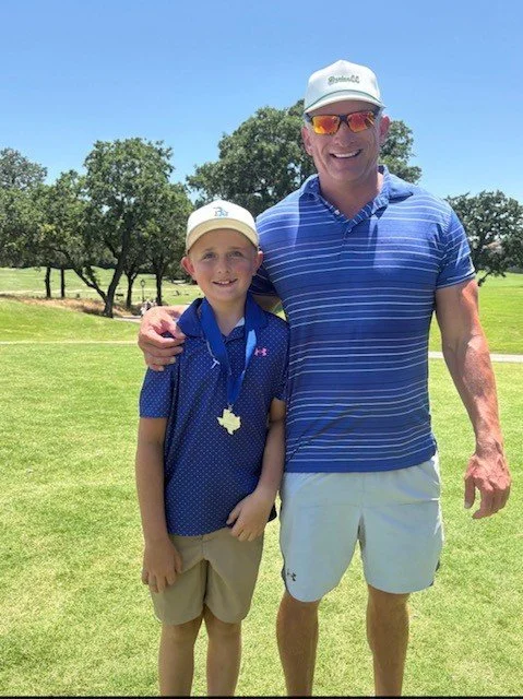 A man and a boy standing together outdoors on a sunny day, smiling and posing for the photo. The boy is wearing a blue polo shirt with a medal around his neck, and the man is dressed in a blue striped polo, white shorts, sunglasses, and a white cap. There are green trees and a grassy field in the background.