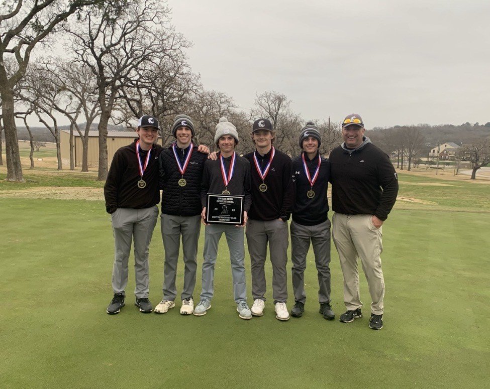 Six people, five young men, and an adult male, standing side by side on a golf course, wearing medals, with a cloudy sky, leafless trees, and some buildings in the background.