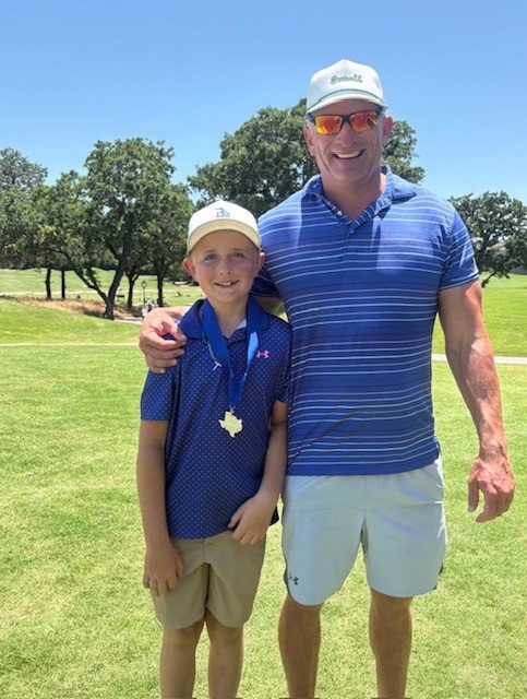 A man and a boy with short blonde hair standing outdoors on a golf course, smiling, with trees in the background. The man is wearing sunglasses, a white cap, a blue striped polo shirt, and white shorts. The boy is wearing a light-colored cap, a blue polo shirt, beige shorts, and has a medal around his neck.