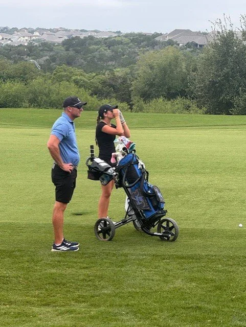 A man and a woman standing on a golf course, with the woman preparing to take a shot and the man watching, with a golf bag on a push cart nearby.
