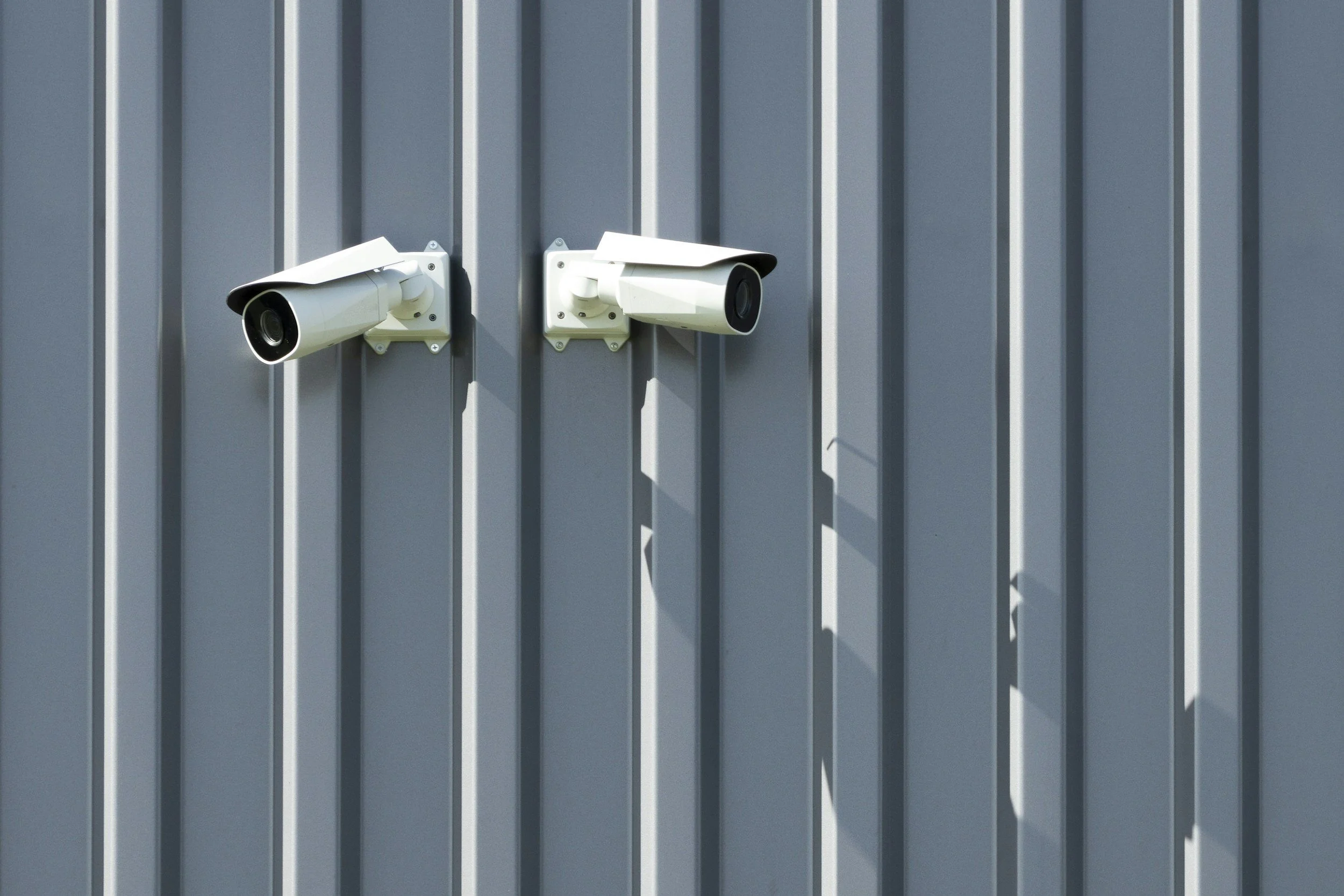 Two white security cameras mounted side by side on a gray corrugated metal wall.
