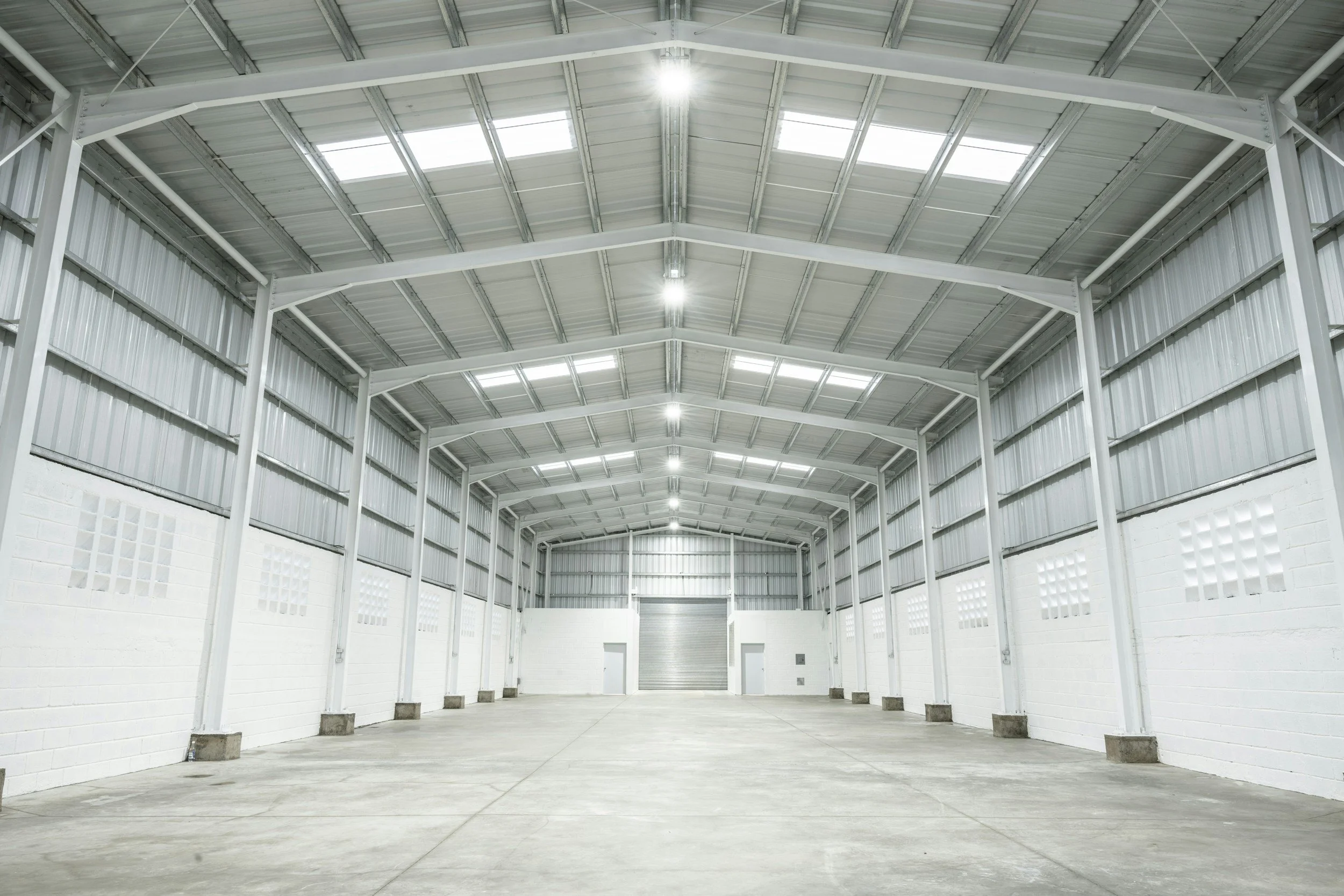 Interior of an empty warehouse with high ceilings, metal roof and wall panels, concrete floor, and bright lighting.