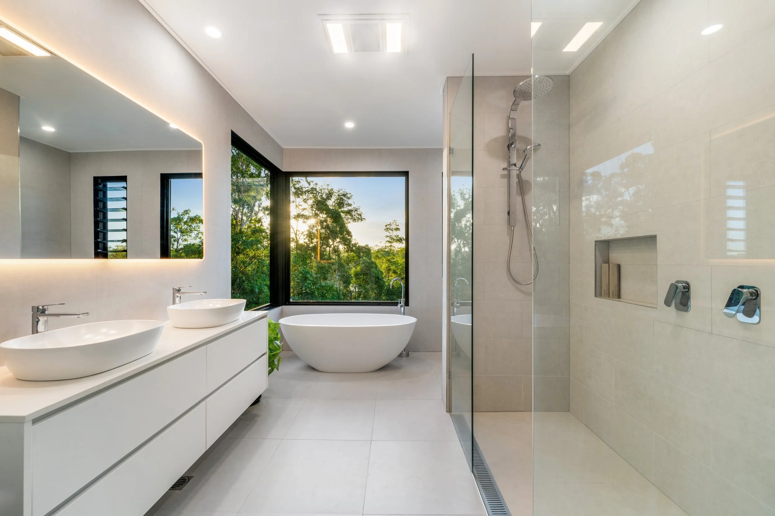 Modern bathroom with large window showing green trees, featuring a freestanding bathtub, double vessel sinks on a white vanity, and a glass walk-in shower with beige tiles and a rainfall shower head.