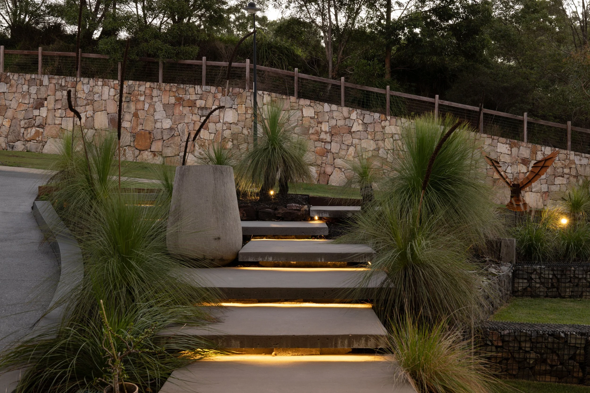 Outdoor stairway with lighting, surrounded by grass, plants, and a stone wall with a wooden fence on top. Trees in the background.
