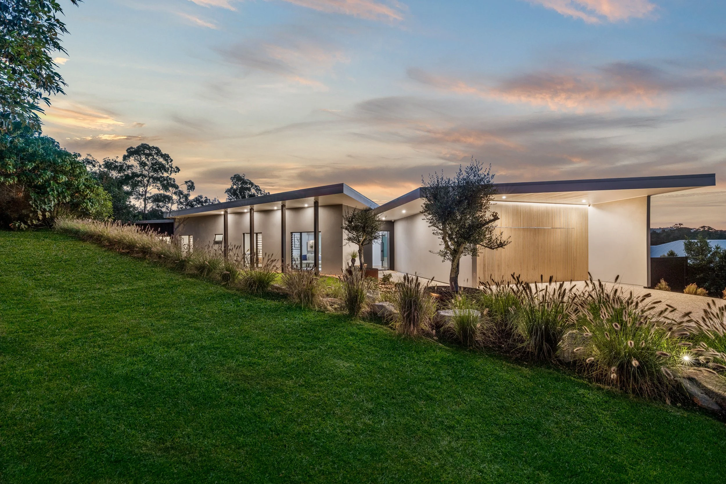 Modern single-story house with large glass windows, surrounded by lush green lawn and trees, under a sky with soft clouds during sunset.