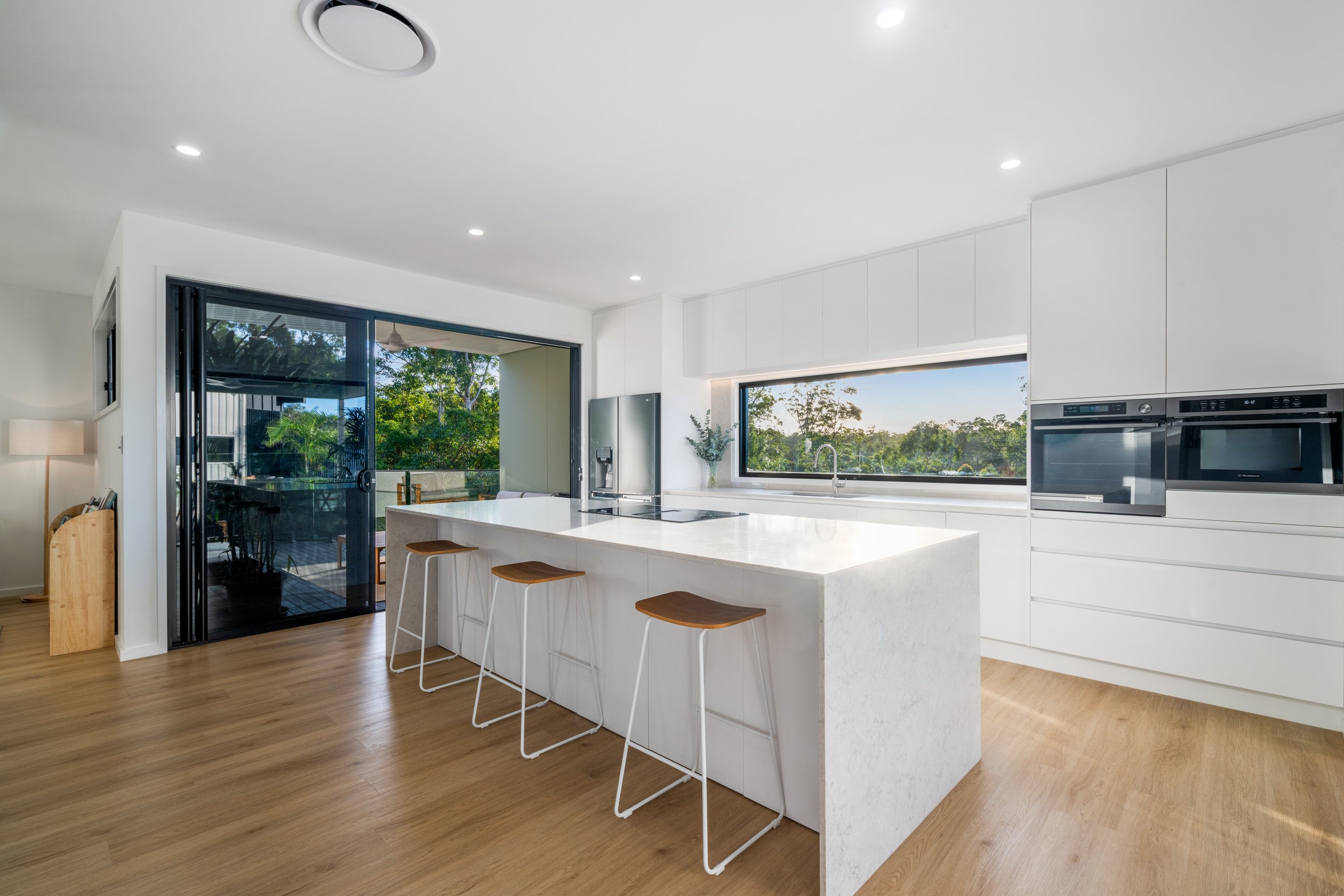 Modern kitchen with white cabinets, a large window, an island with bar stools, and a sliding glass door leading to an outdoor patio with trees.