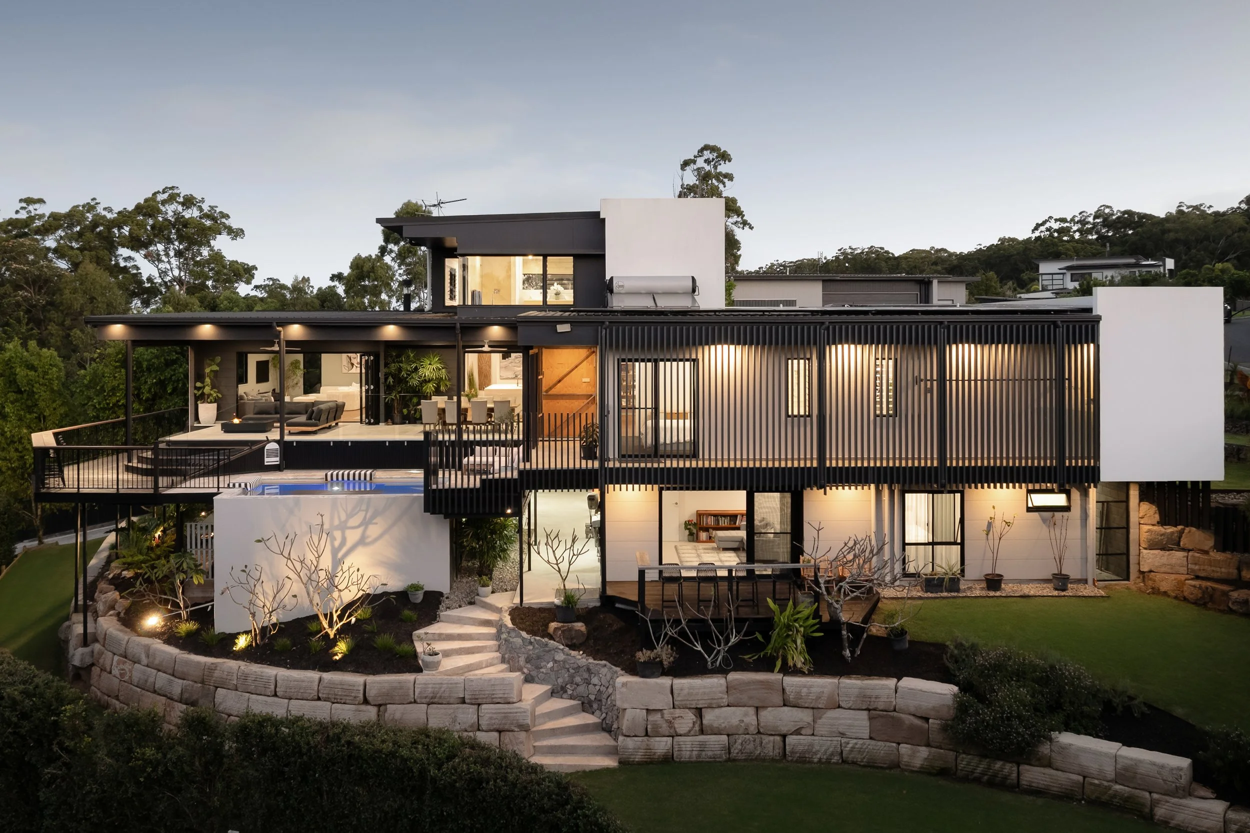 Modern multi-story house with outdoor living area, balcony, and landscaped garden at dusk.