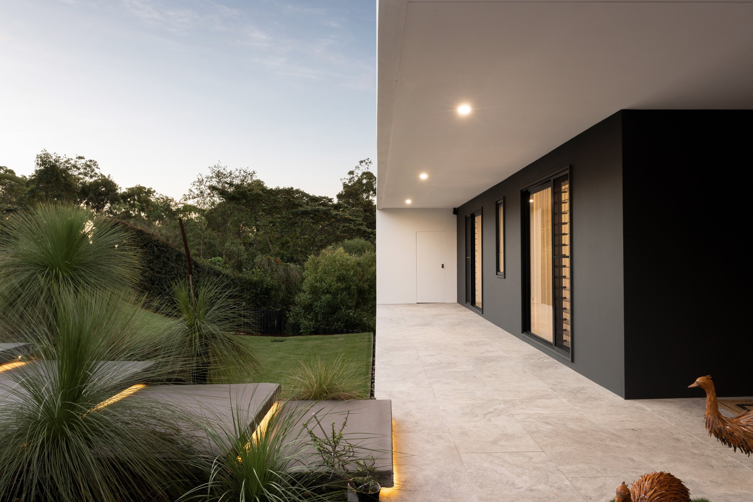 Modern house exterior patio with large sliding glass doors, beige tiled floor, and landscaped garden with shrubs and trees, taken at dusk.