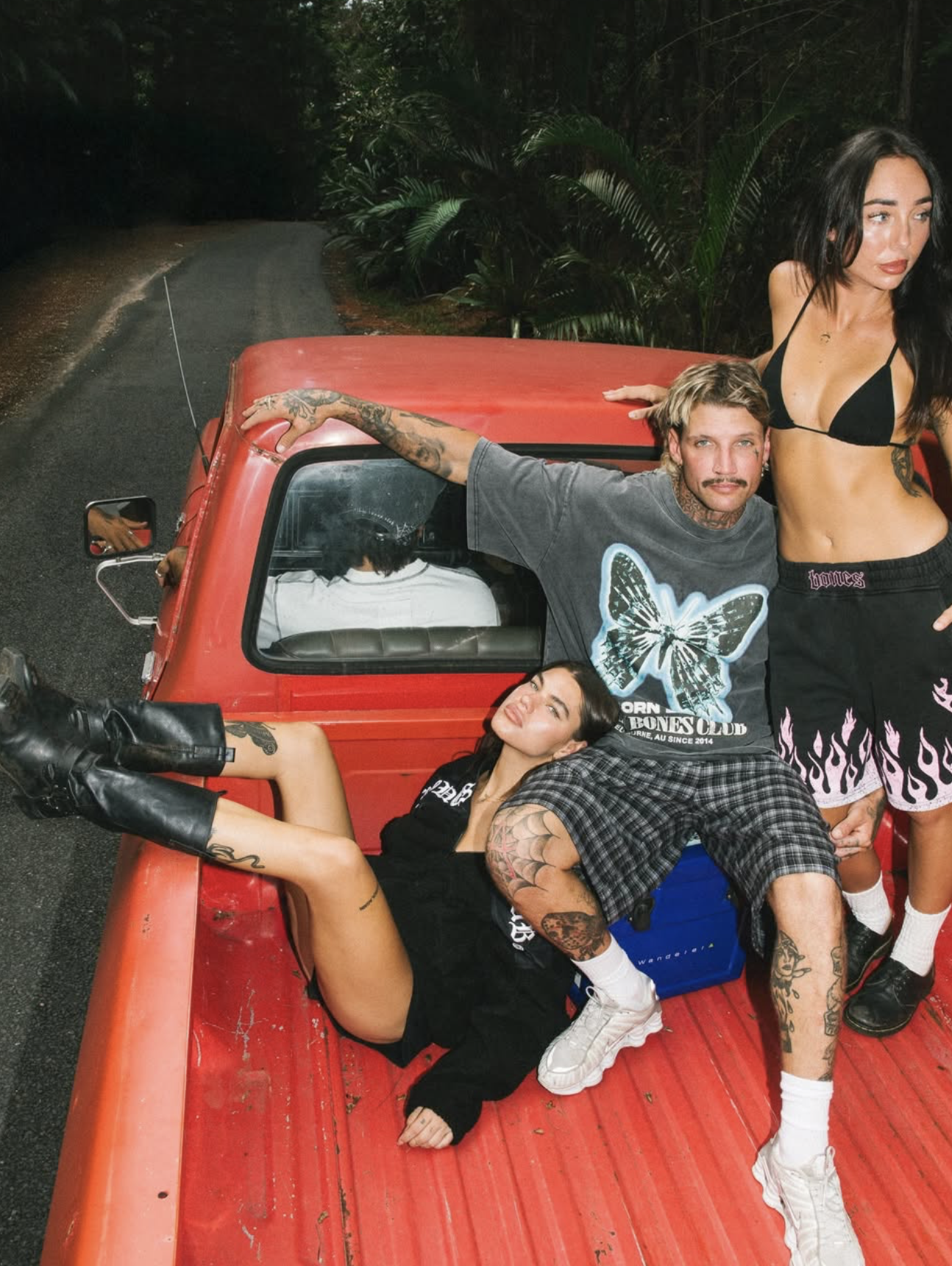 A group of four young adults with tattoos posing on a red pickup truck on a rural road surrounded by trees at night.