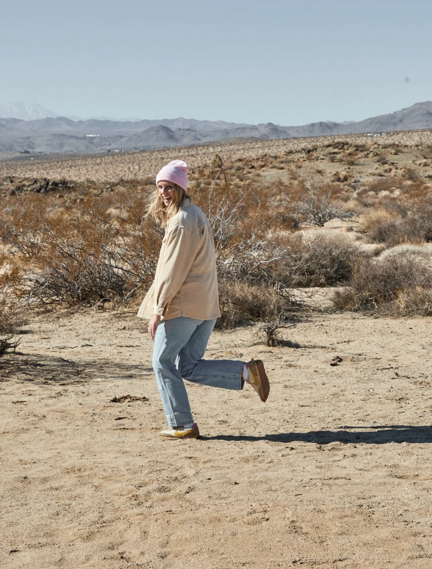 A woman with blonde hair wearing a pink knit hat, beige jacket, gray jeans, and yellow shoes is smiling and playfully kicking one leg up in the air in a desert landscape with shrubs and mountains in the background.