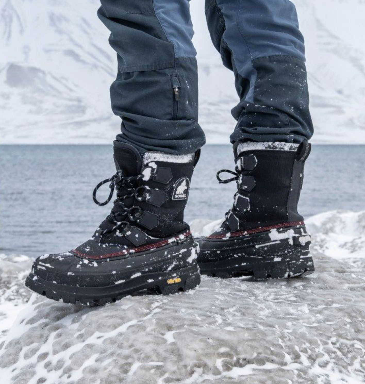 Close-up of person's legs and feet wearing black winter boots and dark blue pants standing on snow-covered ground near water.