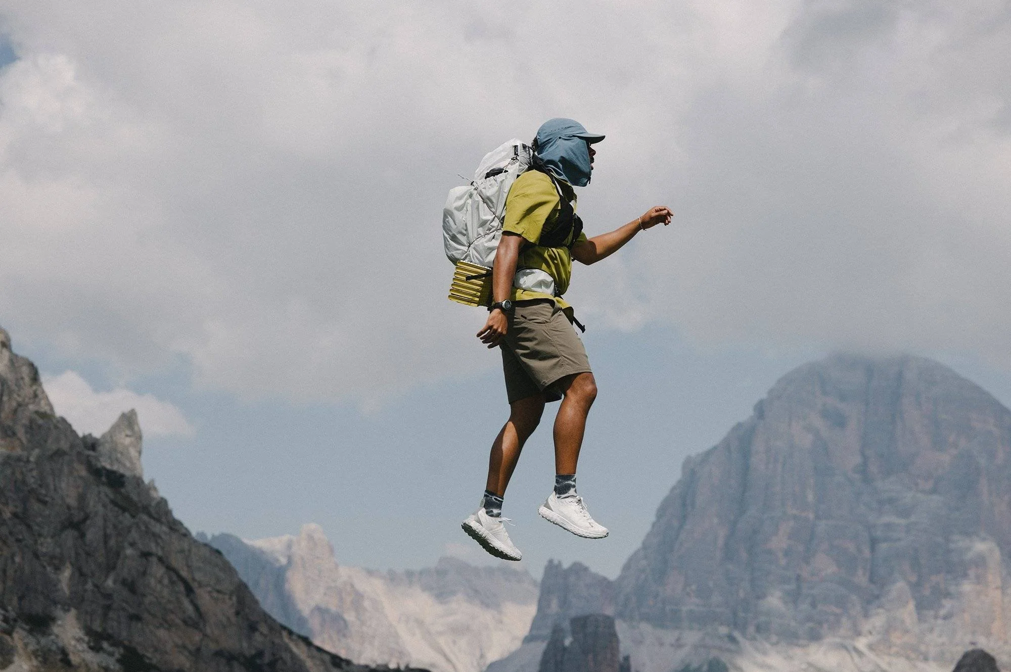 A man wearing hiking gear, including a yellow shirt, gray shorts, and a blue hat, appears to be floating or levitating above a rocky mountain landscape with clouds and distant peaks.