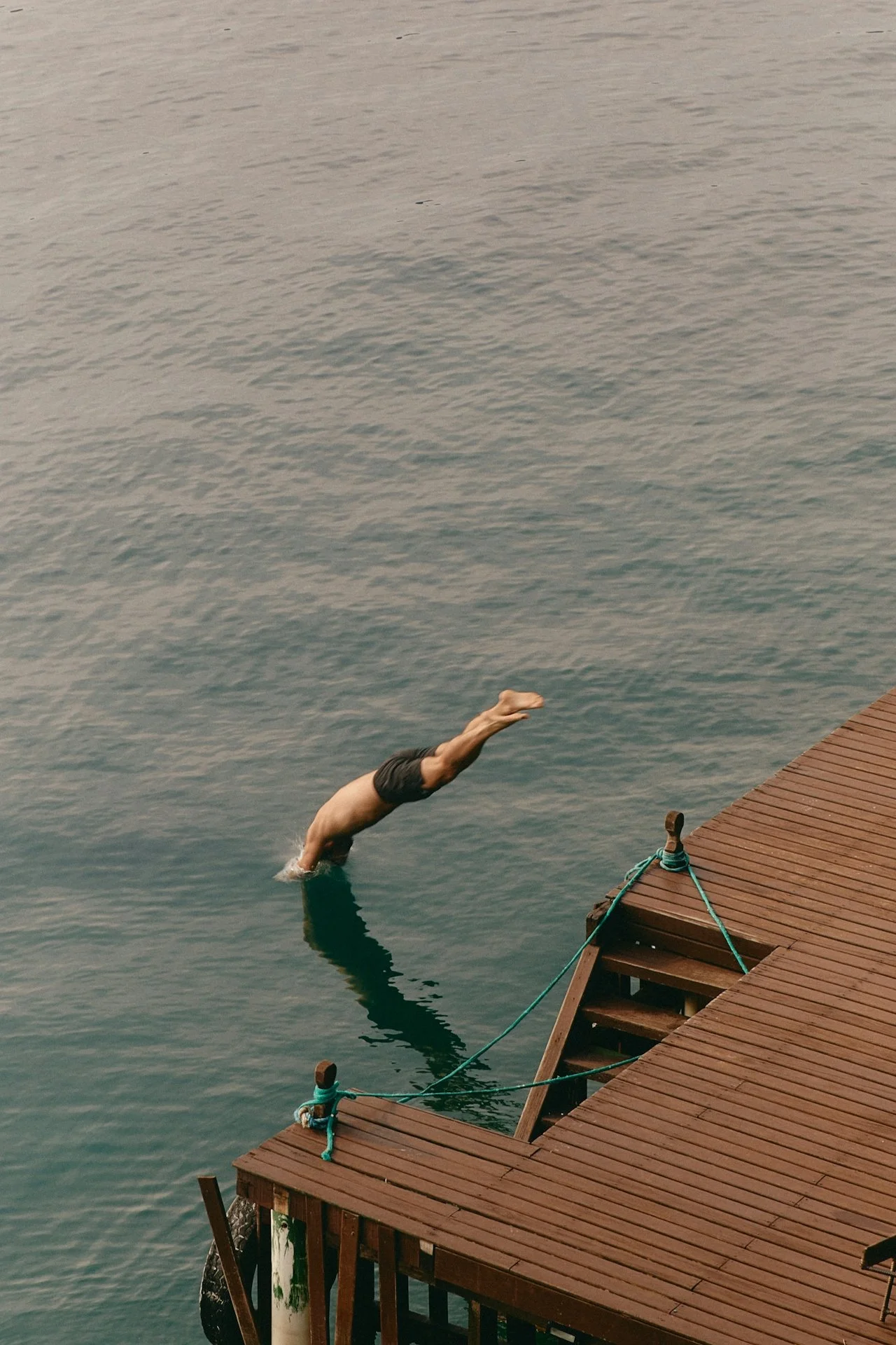 A person is diving into a body of water from a wooden dock, with their body fully extended and head pointed towards the water. The dock is secured with ropes, and the water is calm.