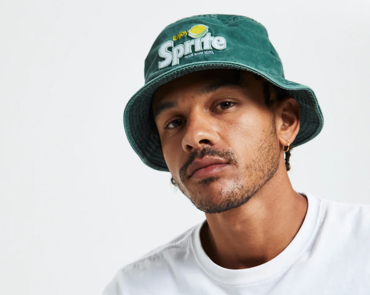 A young man with a serious expression wearing a green bucket hat with a Sprite logo and a white T-shirt, posing against a plain white background.