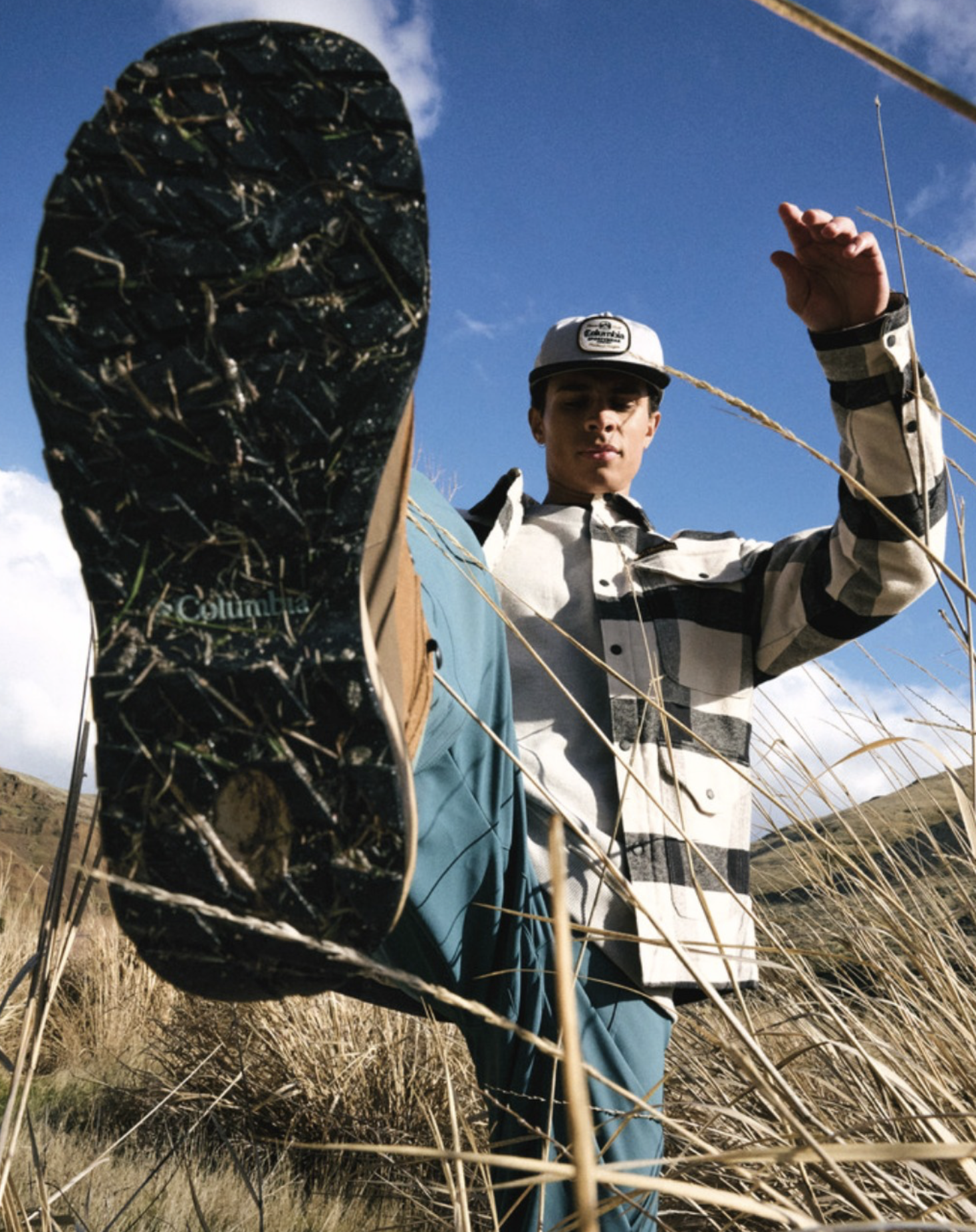 A person wearing a gray and black cap and a checkered shirt standing in a field of dry grass, with their foot closest to the camera, wearing a black and green Columbia shoe, raised up to show the sole. The sky is blue with some clouds.