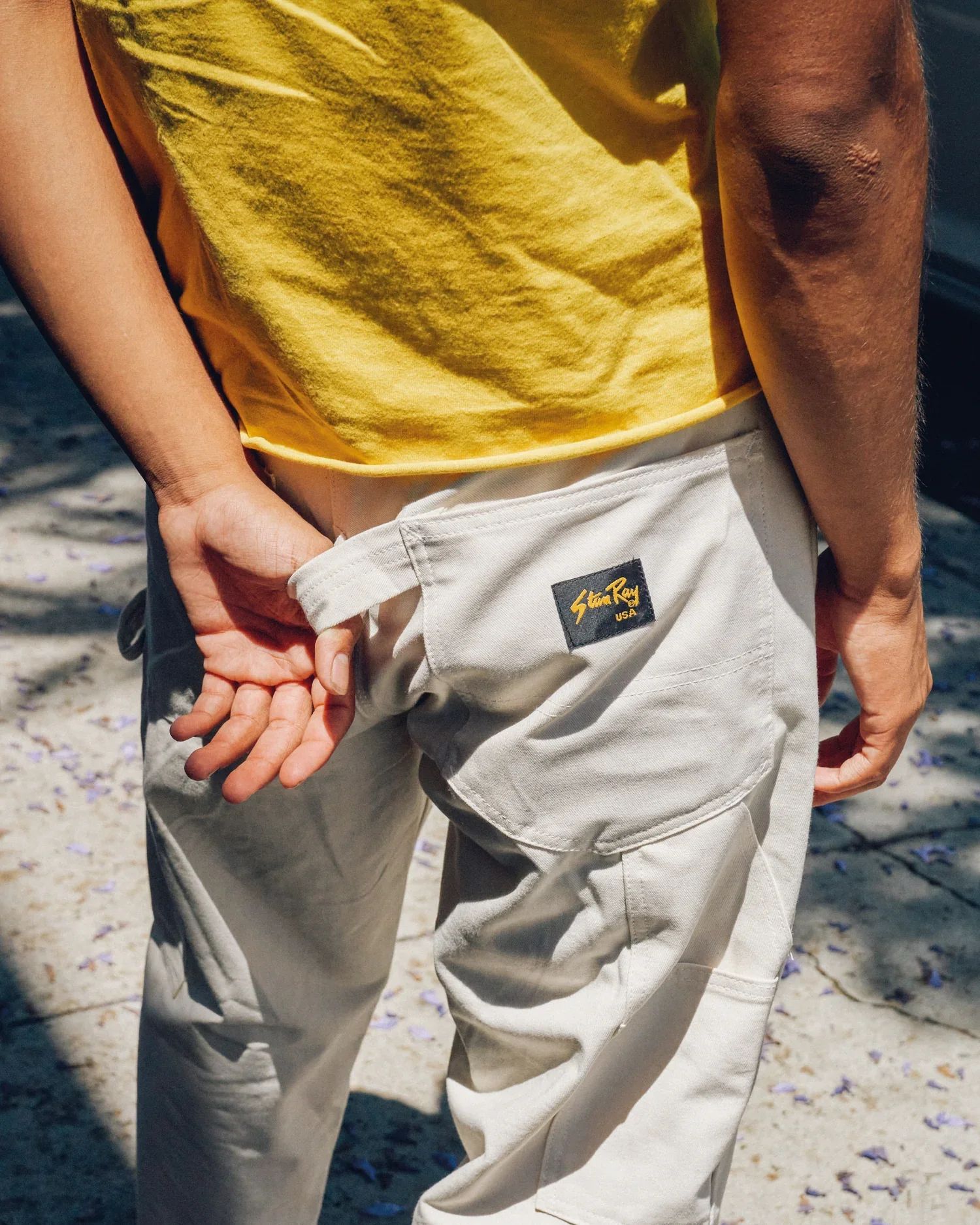 Close-up of a person wearing a yellow shirt and white pants holding hands with another person on a sandy surface with purple flower petals.