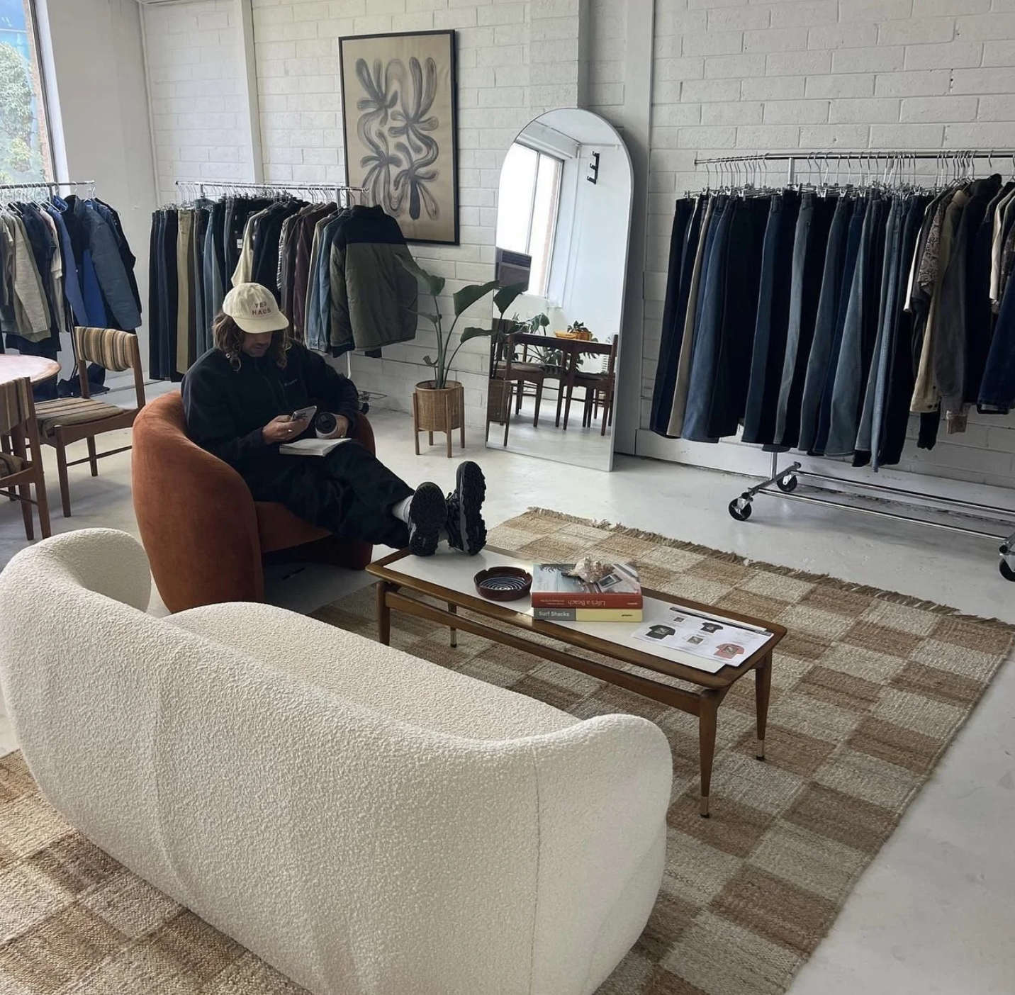 A person sitting on a burnt orange armchair, looking at their phone, in a fashion boutique with clothing racks, a large mirror, artwork, a white textured sofa, and a coffee table with magazines.