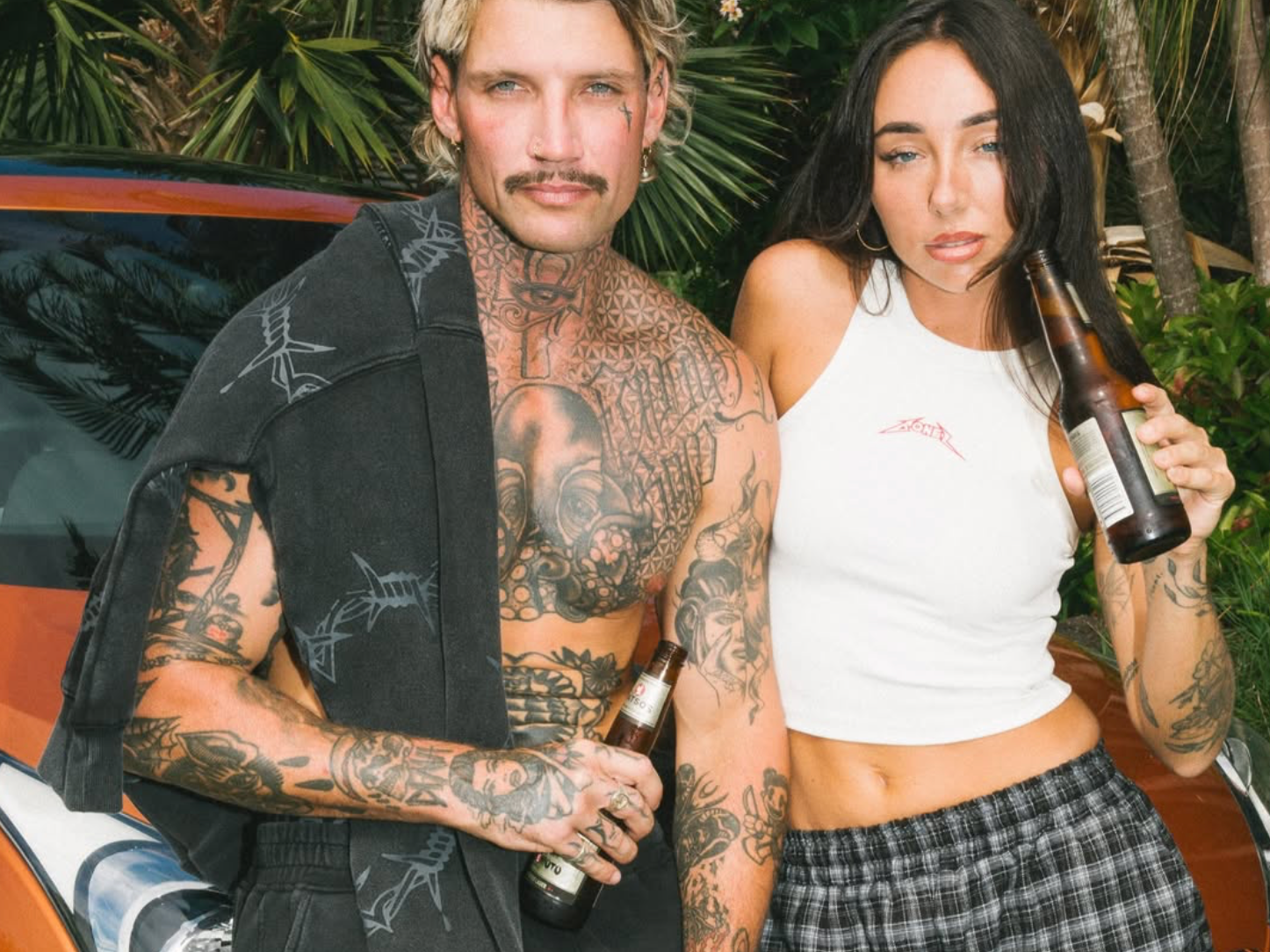 A shirtless man with tattoos and a woman in a white tank top holding beer bottles standing outdoors near a car and green foliage.