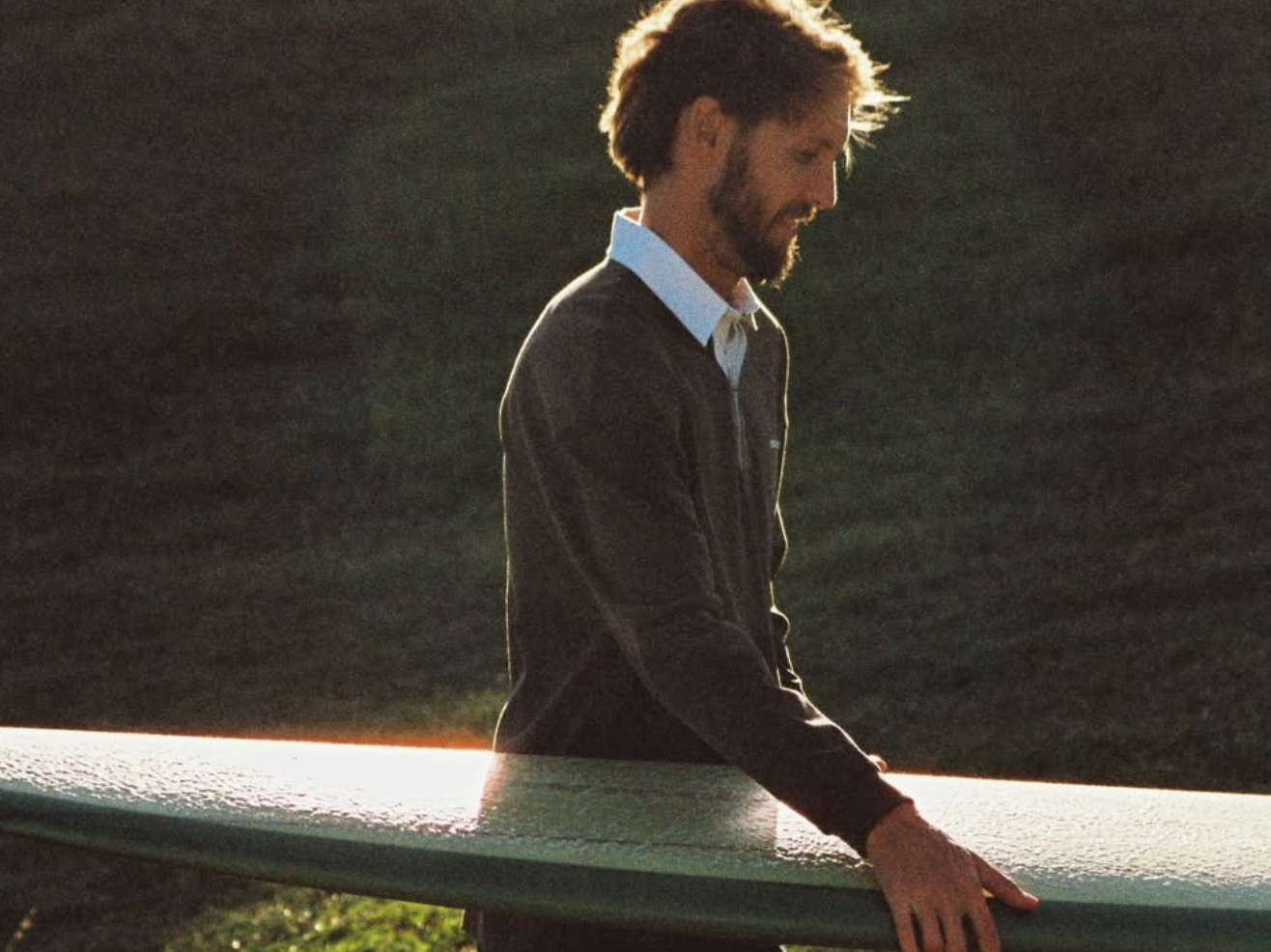 A man with brown hair and a beard, wearing a dark jacket and white shirt, standing outdoors during sunset, holding a surfboard horizontally with both hands.